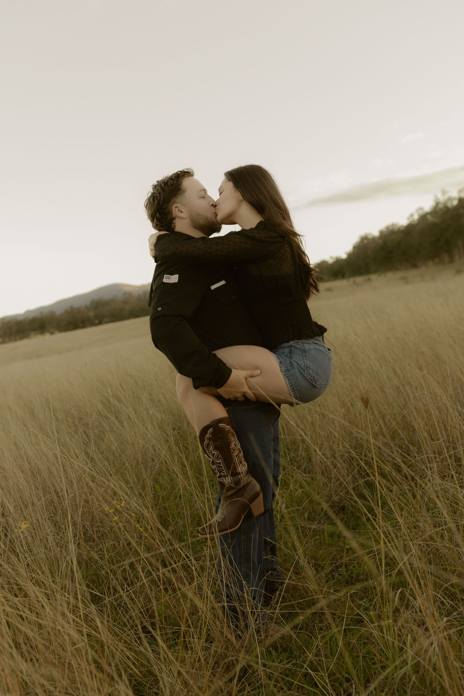 A man lifts and kisses a woman in denim shorts and cowboy boots in a vast golden grass field at dusk.
