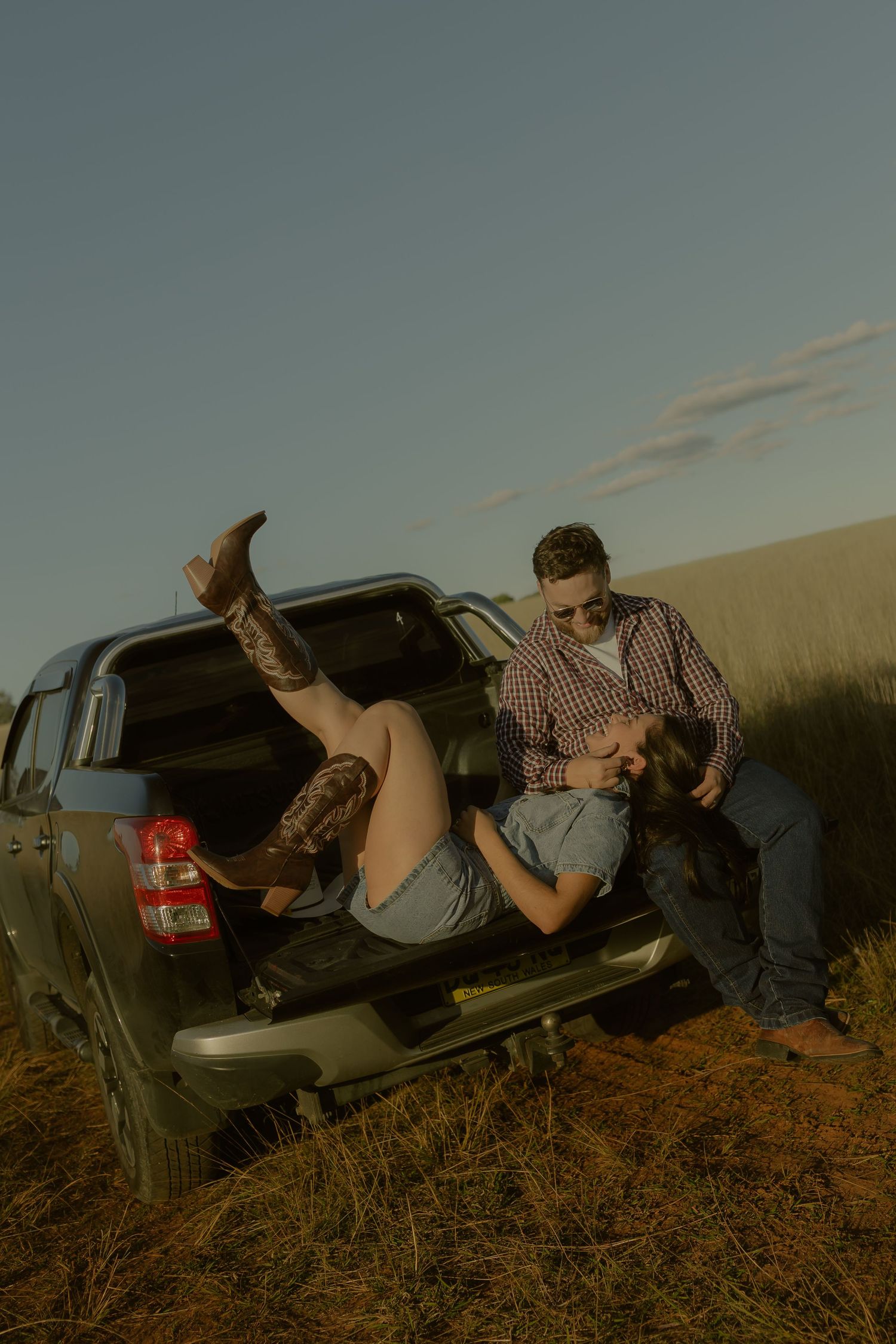 Romantic couple relaxing in the back of a pickup truck at sunset in a rural field, woman wearing cowboy boots.