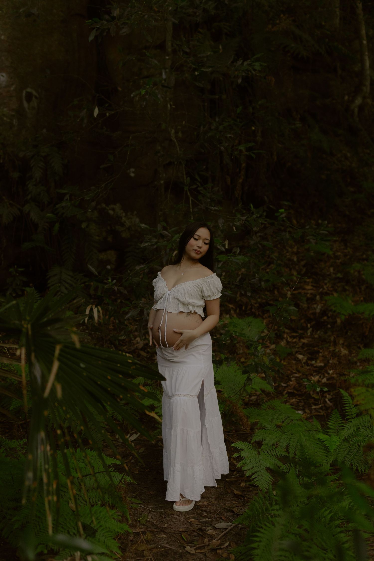 Pregnant woman in white dress standing on forest path surrounded by lush green ferns and trees.