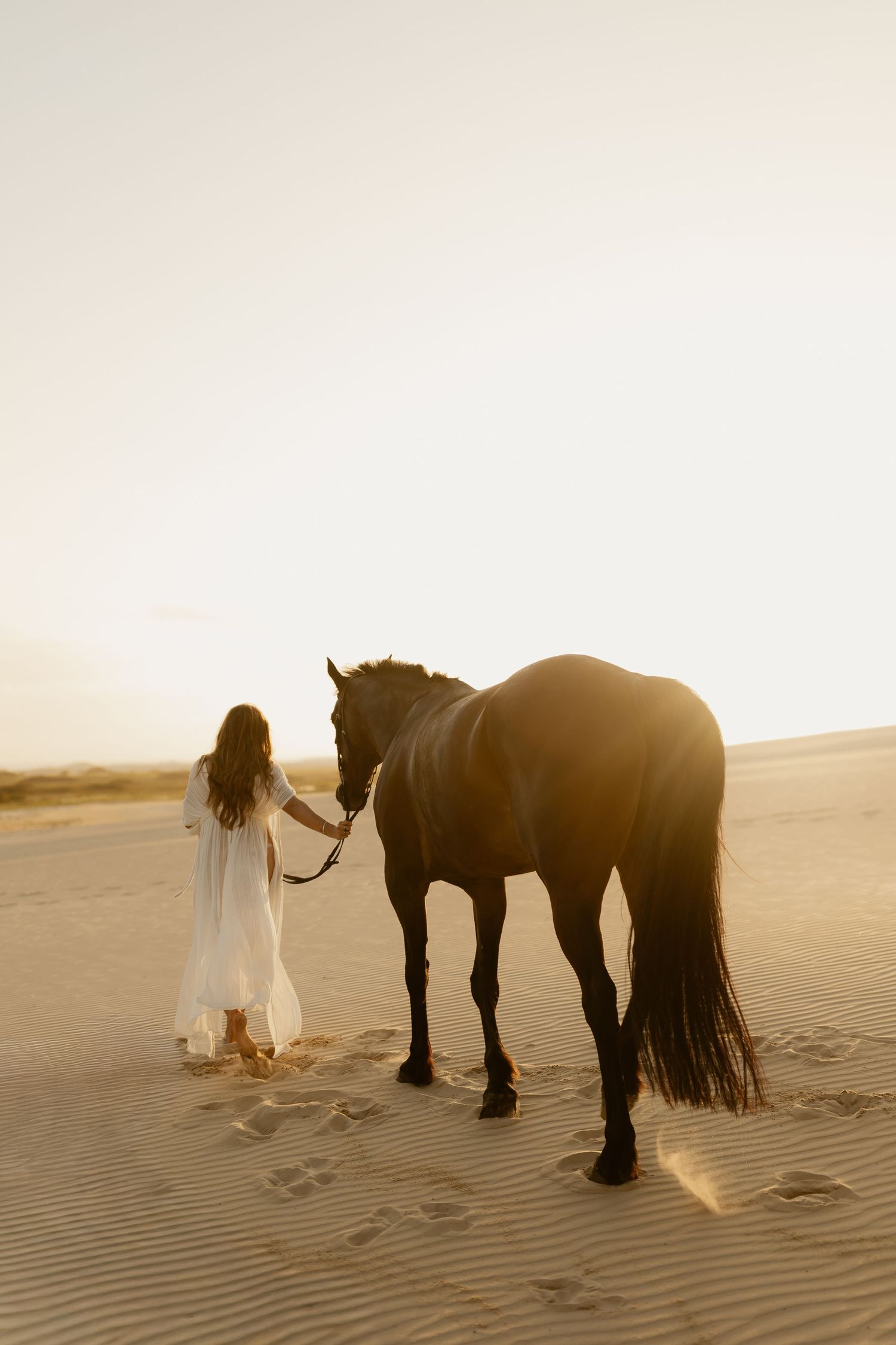 A silhouetted figure in a flowing dress stands beside a horse on sand dunes during golden hour sunset.