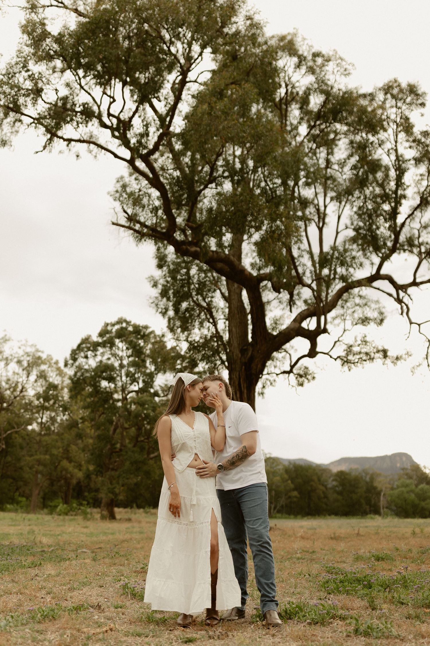 Romantic couple shares intimate moments in a rustic outdoor setting with rolling hills and tall trees in the background.