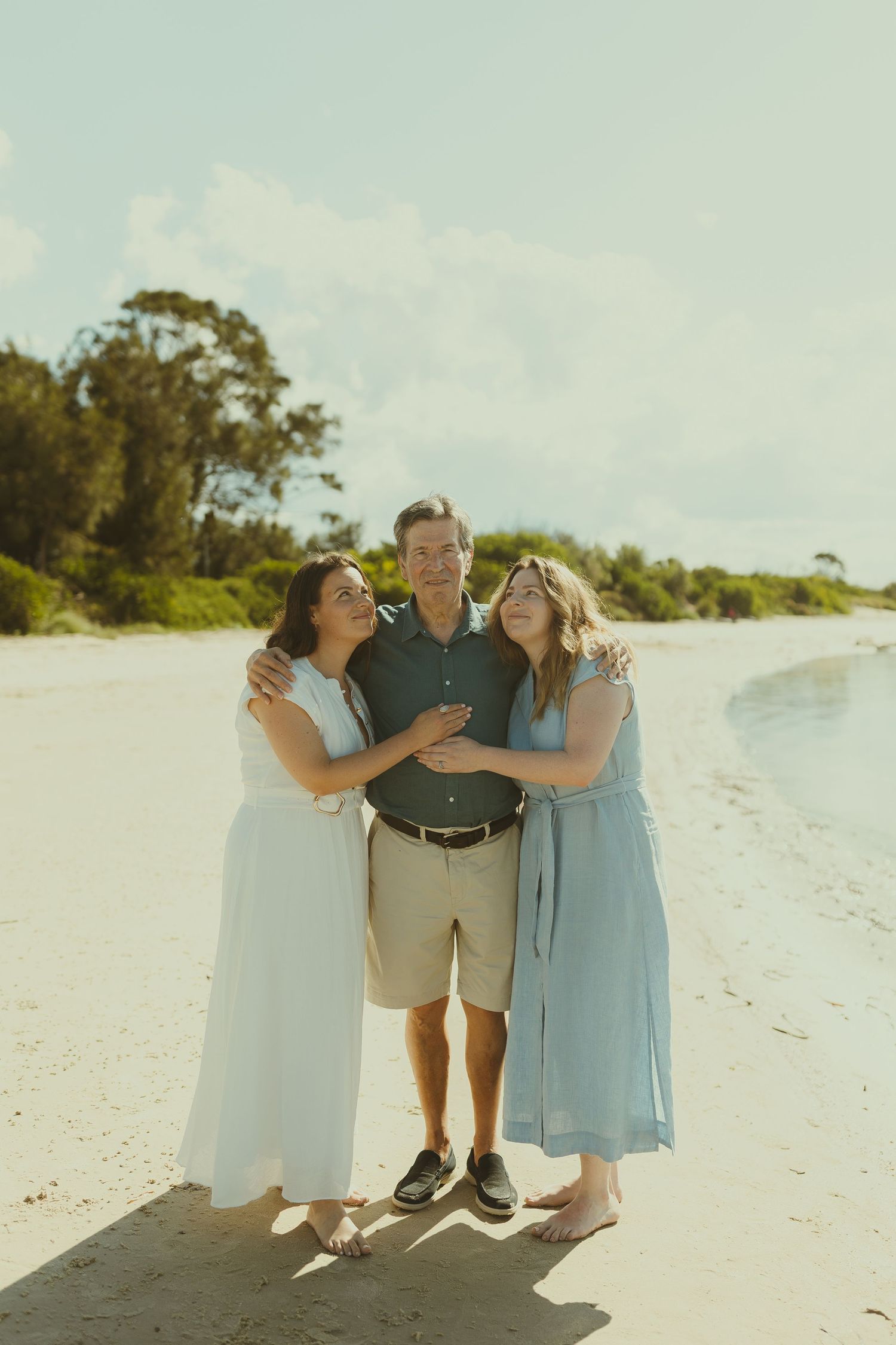 Three people embrace and laugh together while standing on a pristine white sandy beach.