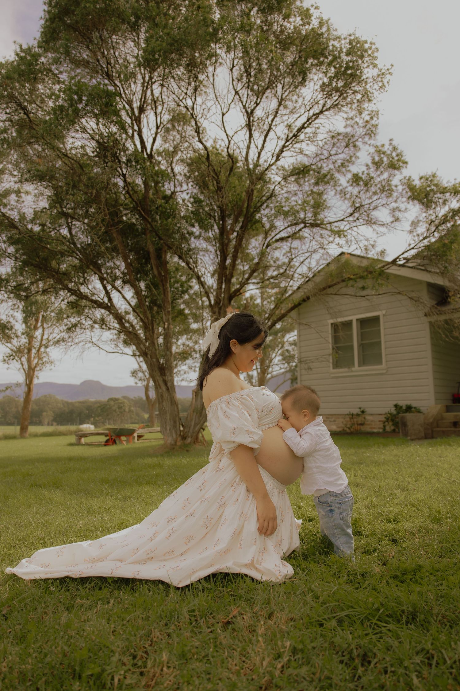 Pregnant woman in white floral dress kneels as toddler touches her baby bump outdoors near farmhouse.