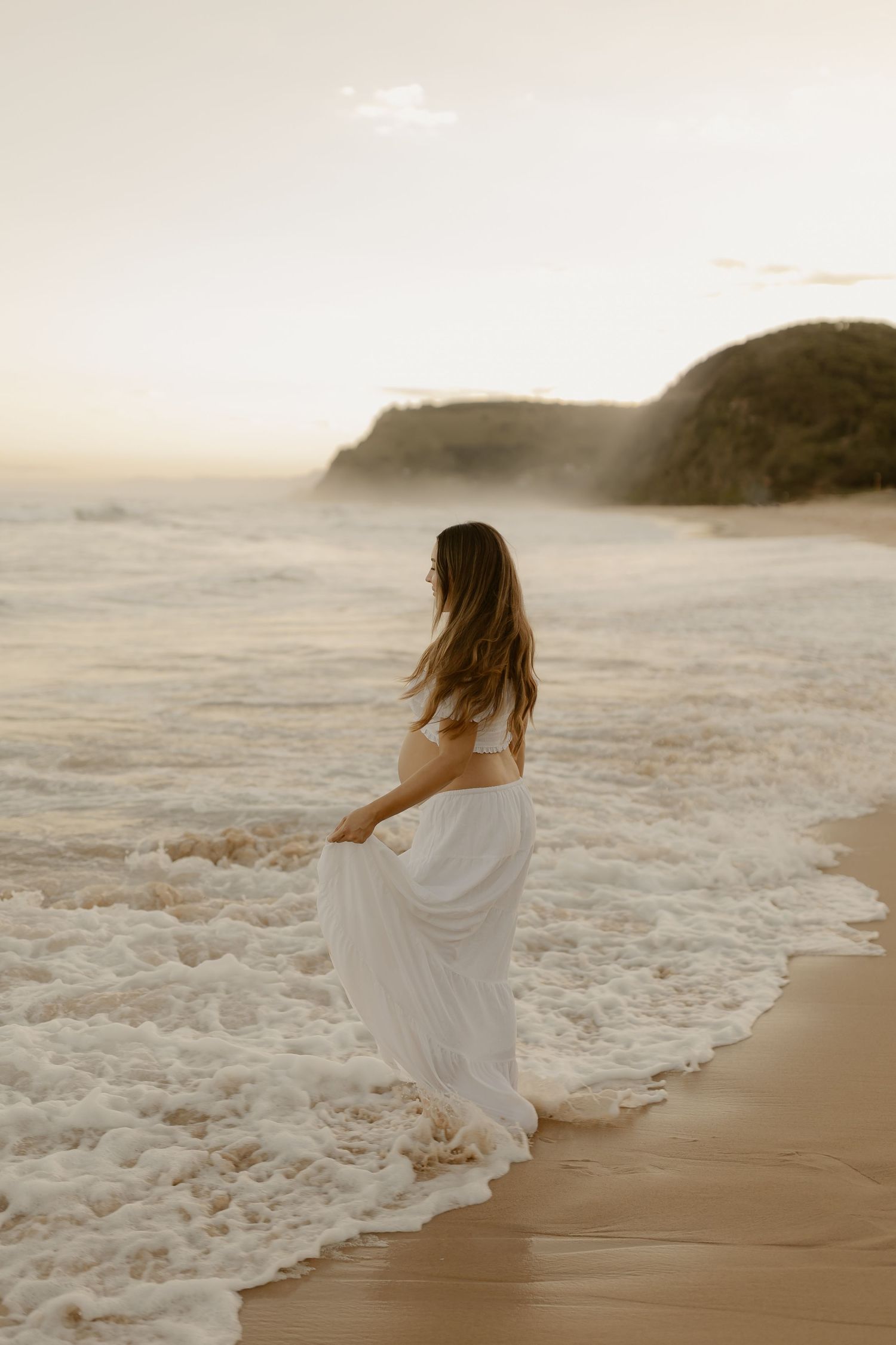 A figure in a flowing white dress dances along a misty beach shoreline at sunset.