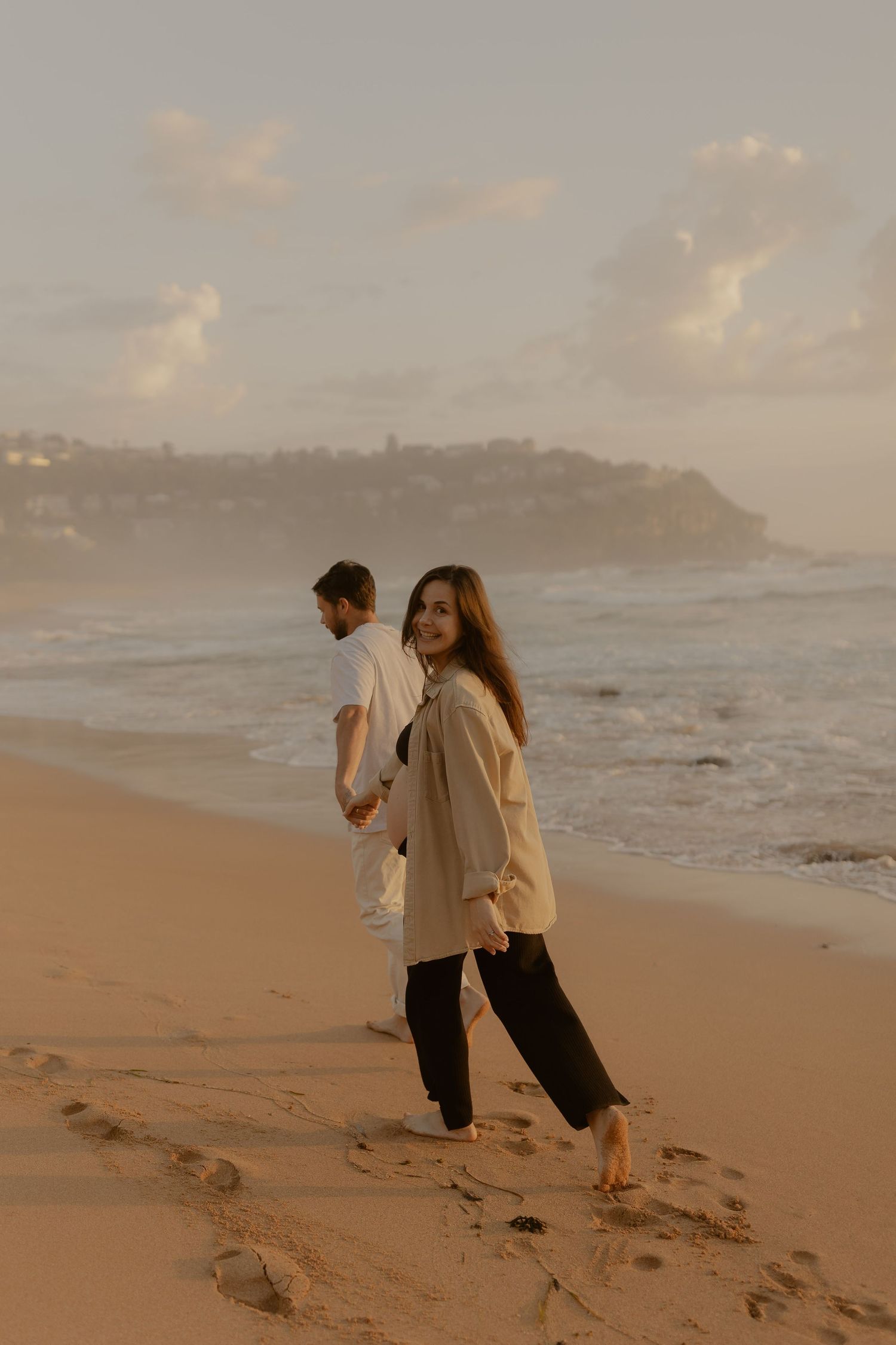 A couple walks along a sandy beach at sunset, leaving footprints in their wake as waves crash nearby.