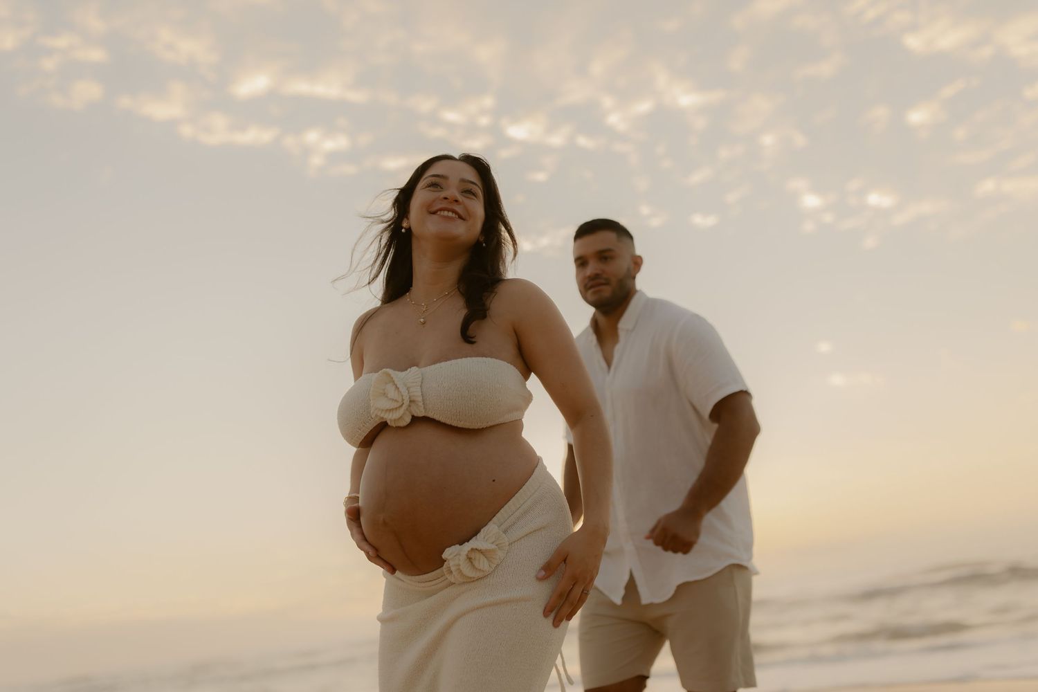 A couple embraces on a beach during sunset while posing for romantic maternity photos.