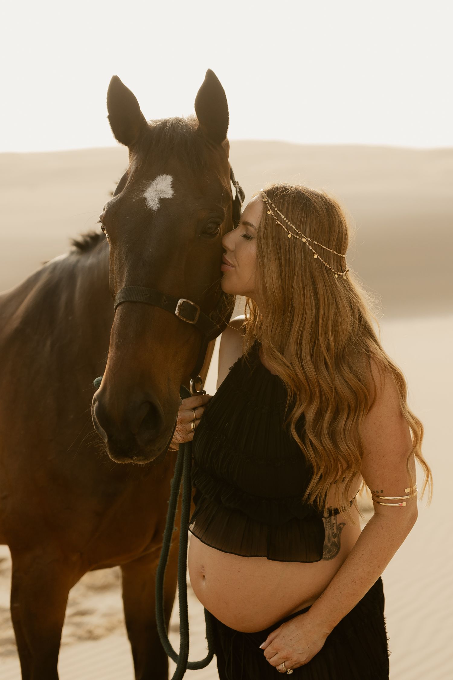 A black horse and woman in a flowing dress share an intimate moment on a sandy beach at sunset.