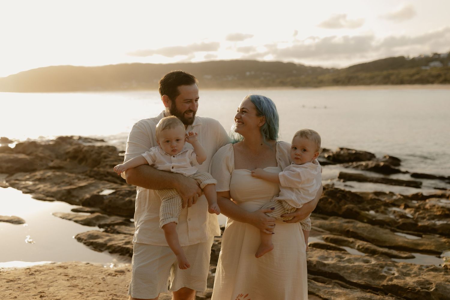 A family dressed in white embraces on a rocky coastline during golden hour.