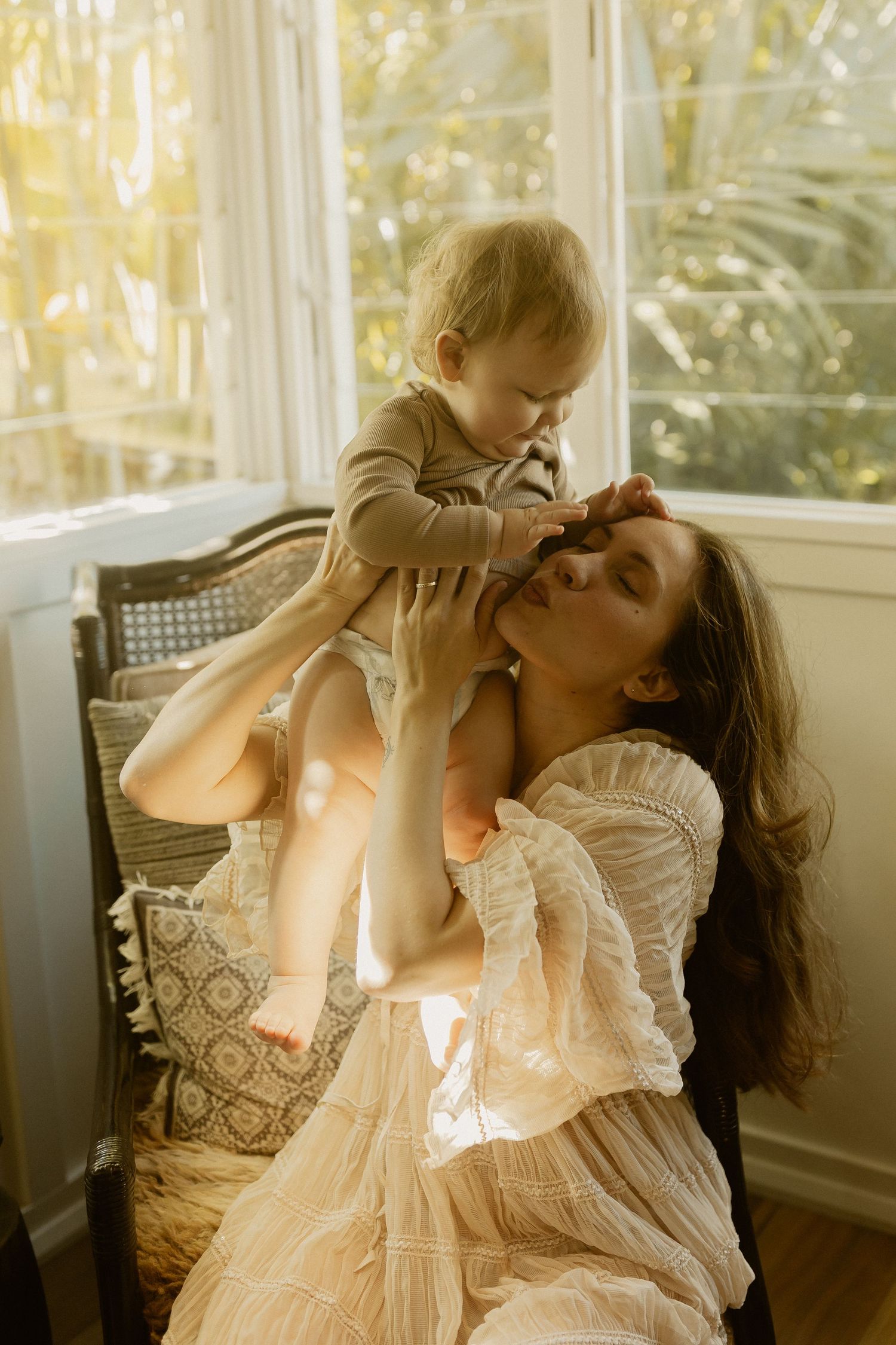 Parent and baby playing together by sunny window in flowing white dress.