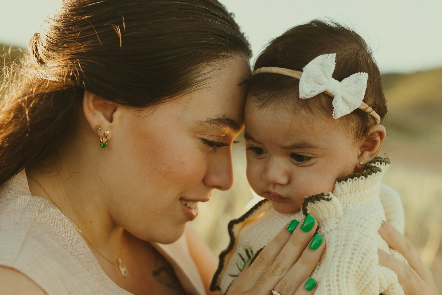 A tender moment captured between a parent and baby wearing a white bow headband in warm sunlight.