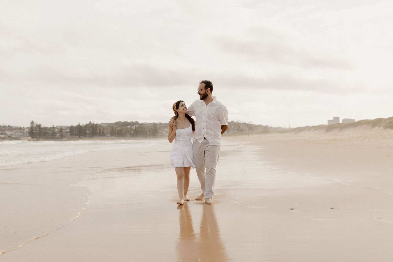 A couple walks together on a serene beach at sunset, leaving footprints in the wet sand.
