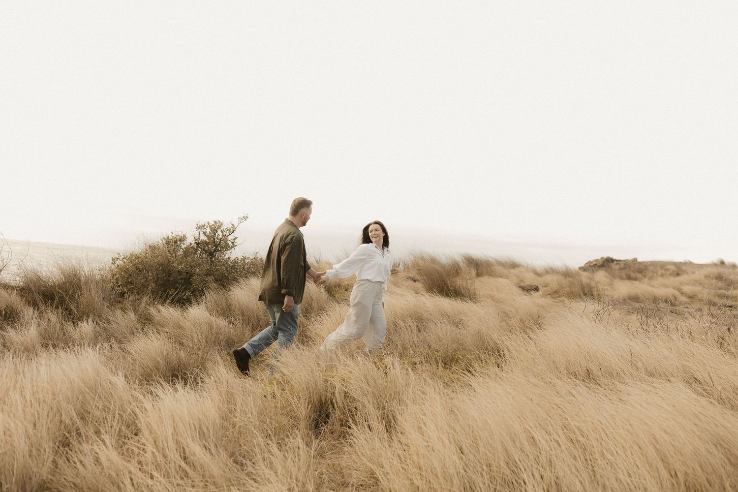 A couple walks together through tall golden grass on a scenic hillside during sunset.