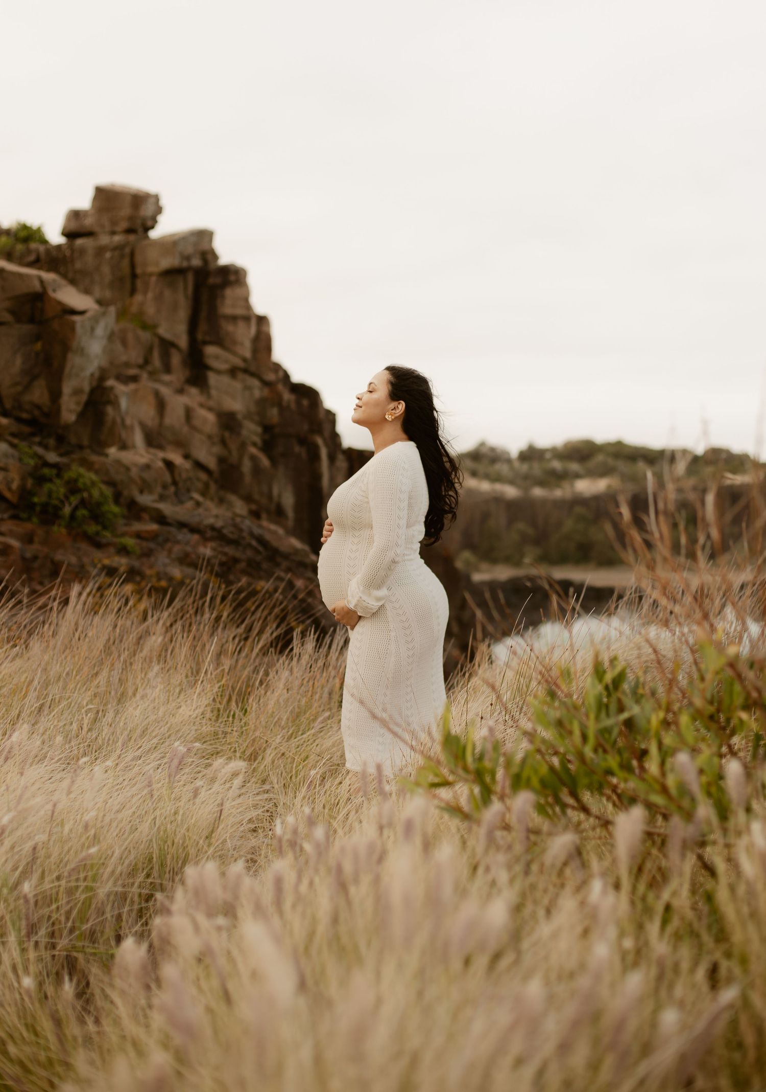 A person in a flowing white dress stands gracefully in a field of tall grass against a rocky landscape backdrop.