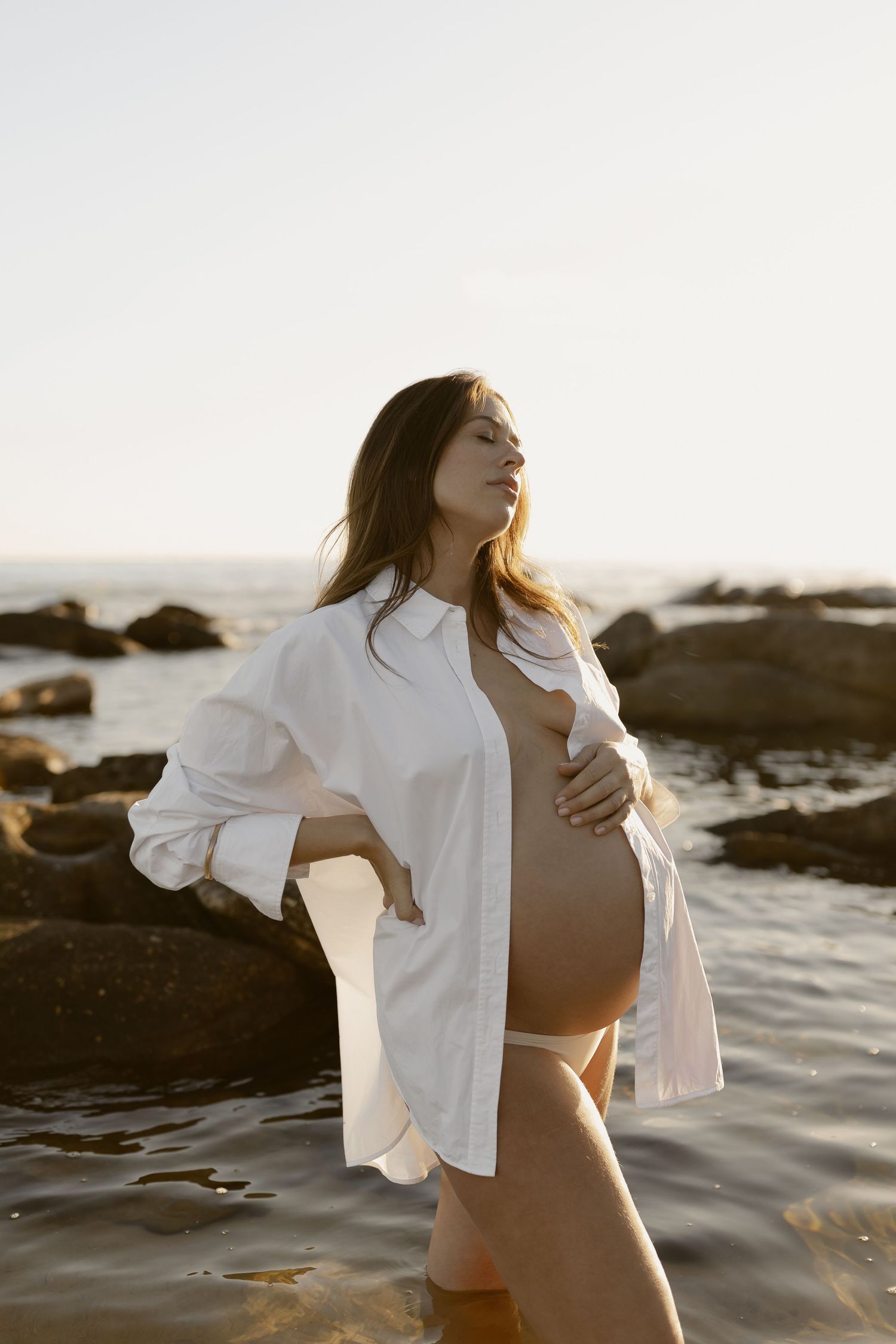 Maternity photo of a person in white shirt standing by ocean rocks at sunset.