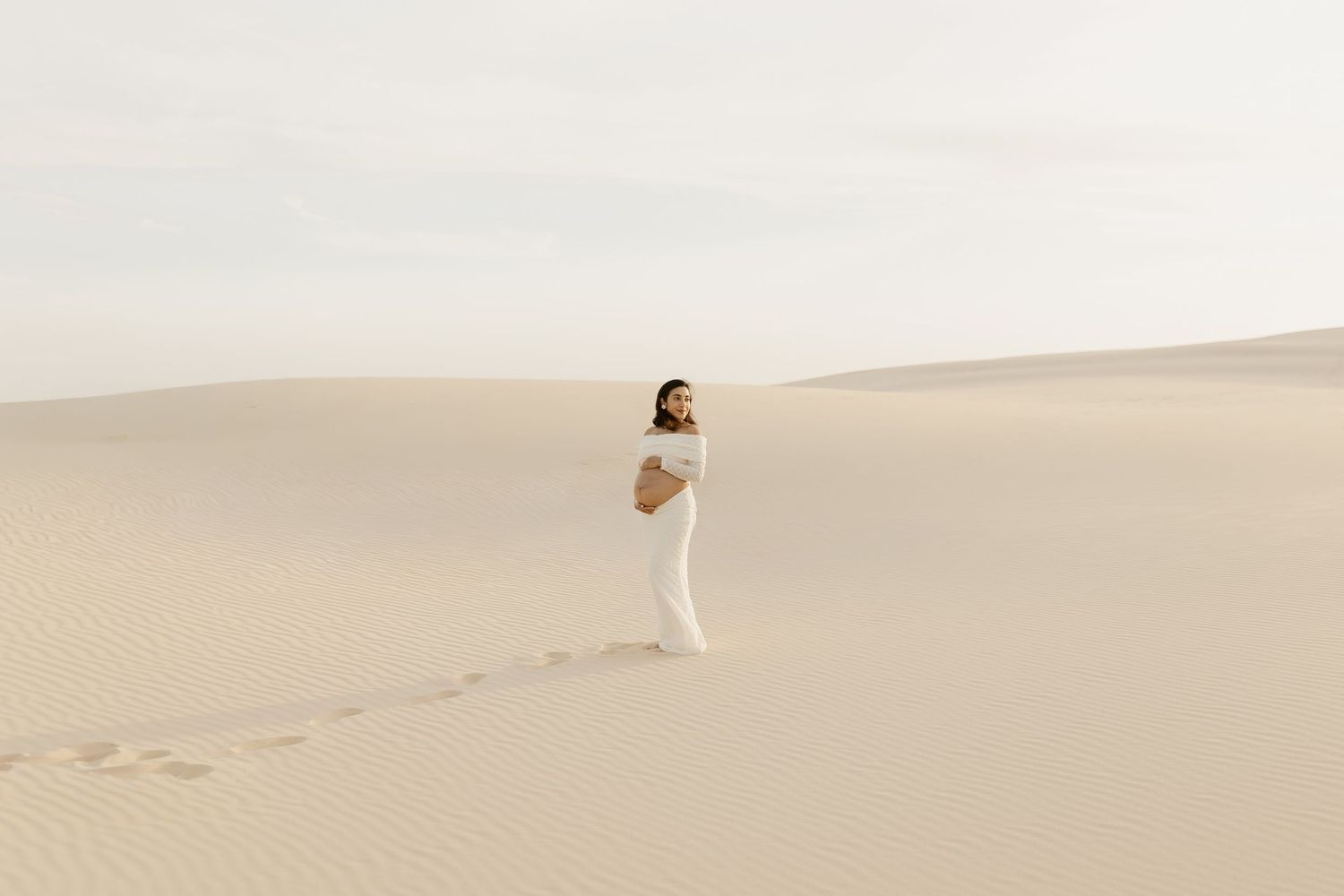 A person in a white dress stands gracefully in the vast sandy dunes of a desert landscape at sunset.