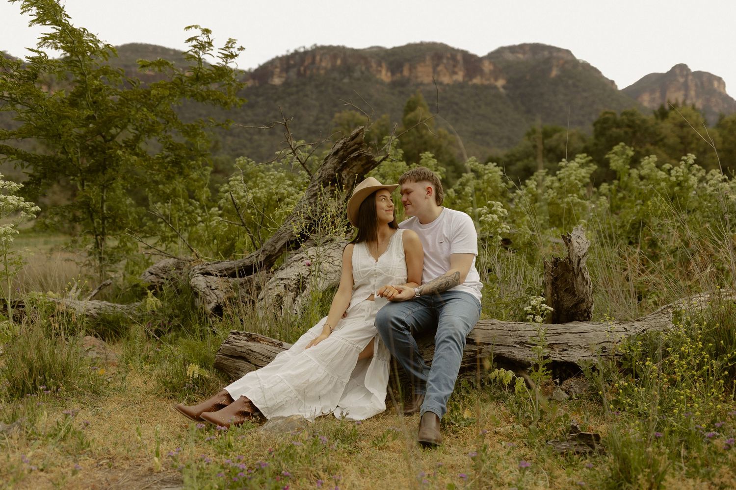 Romantic couple in white outfits sitting together on grassy hillside with mountain landscape in background.
