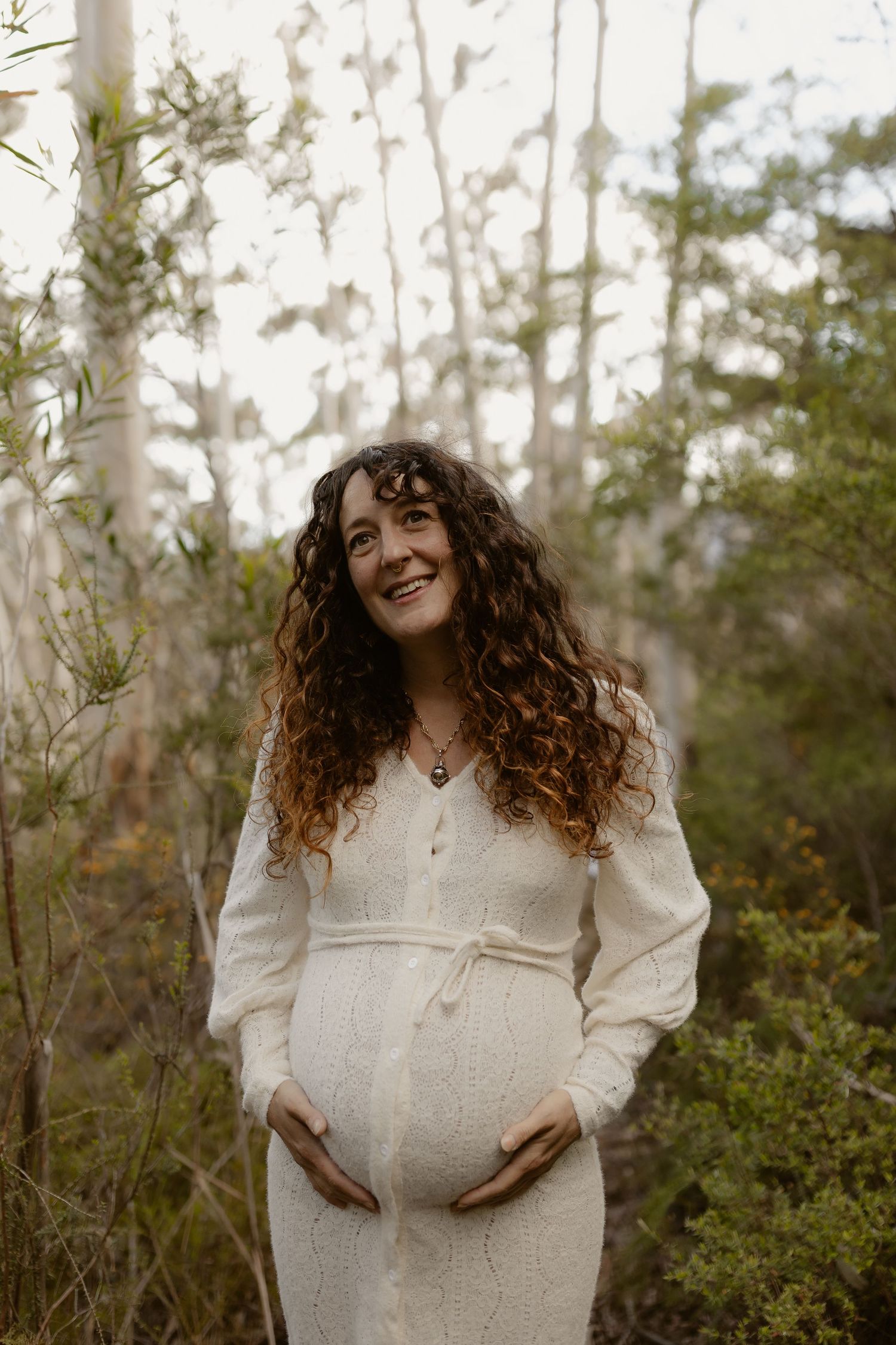 A woman in a white sweater stands in a natural outdoor setting with trees and greenery in the background.