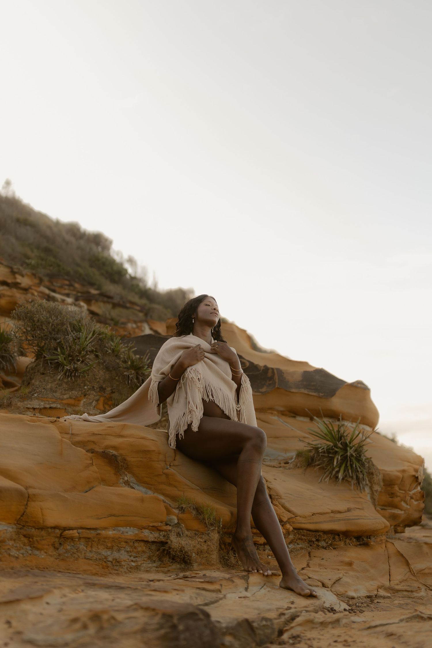 Woman in fringe shawl sitting on sandstone rocks at golden hour, head tilted back.