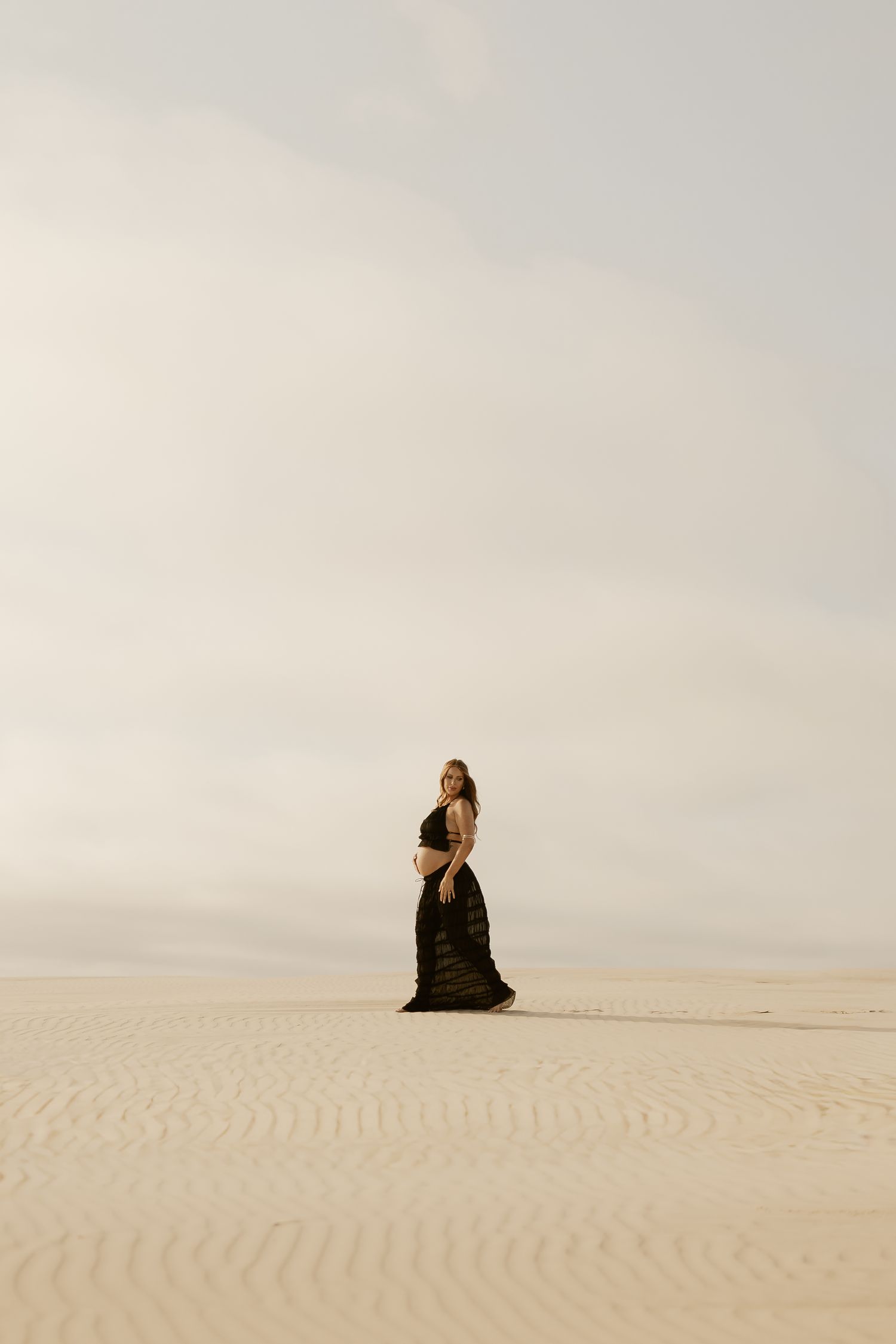 A pregnant woman in a black dress stands alone on vast white sand dunes under a cloudy sky.