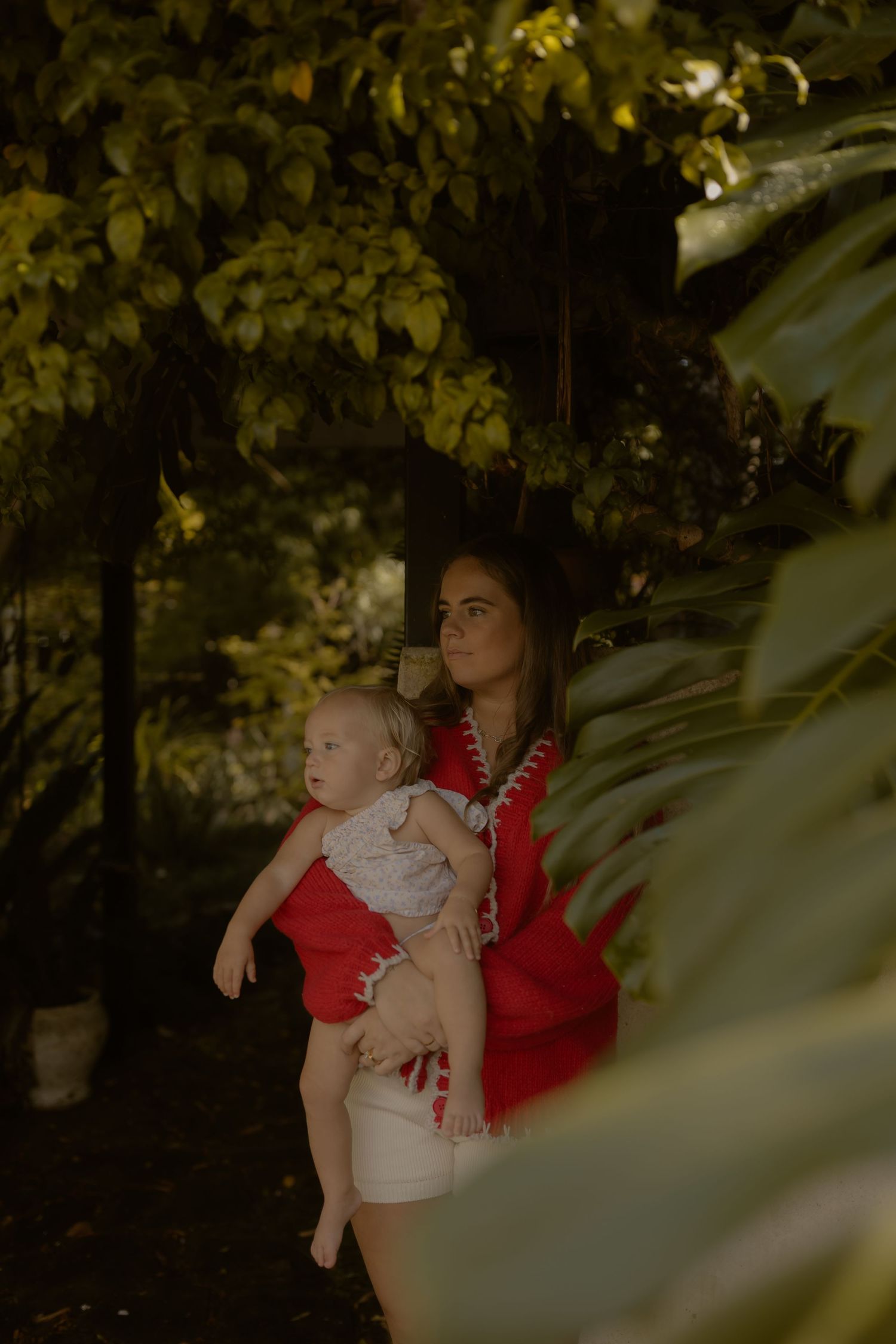 Mother in red fringed top holding baby outdoors among lush green tropical garden foliage.