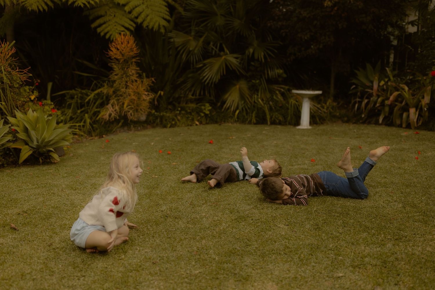 Three young children playing on lush green grass in a garden surrounded by tropical plants and palm trees.