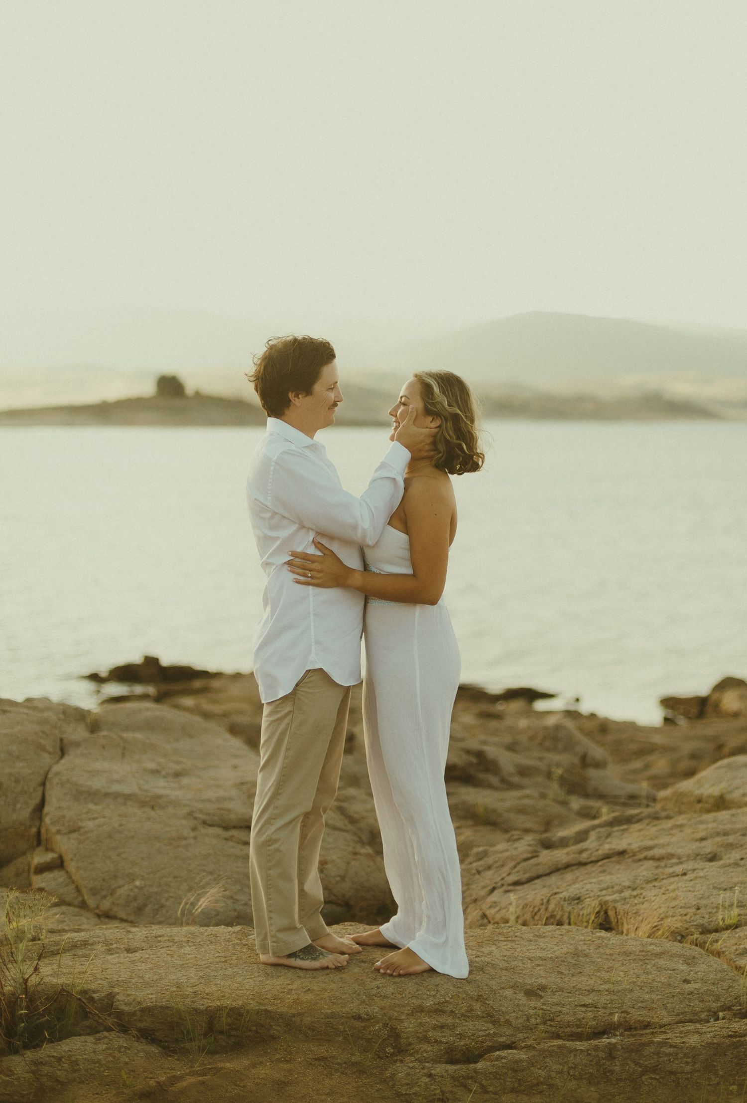 A couple in white and beige outfits embrace romantically on coastal rocks at sunset with ocean and mountains in background.