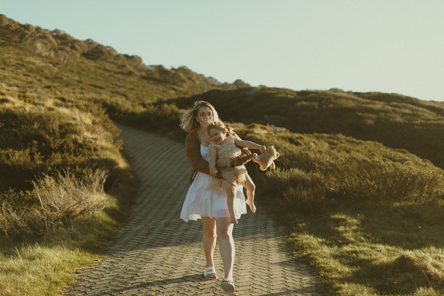 A person in a white dress dances along a winding dirt path through golden grass-covered hills in warm sunlight.