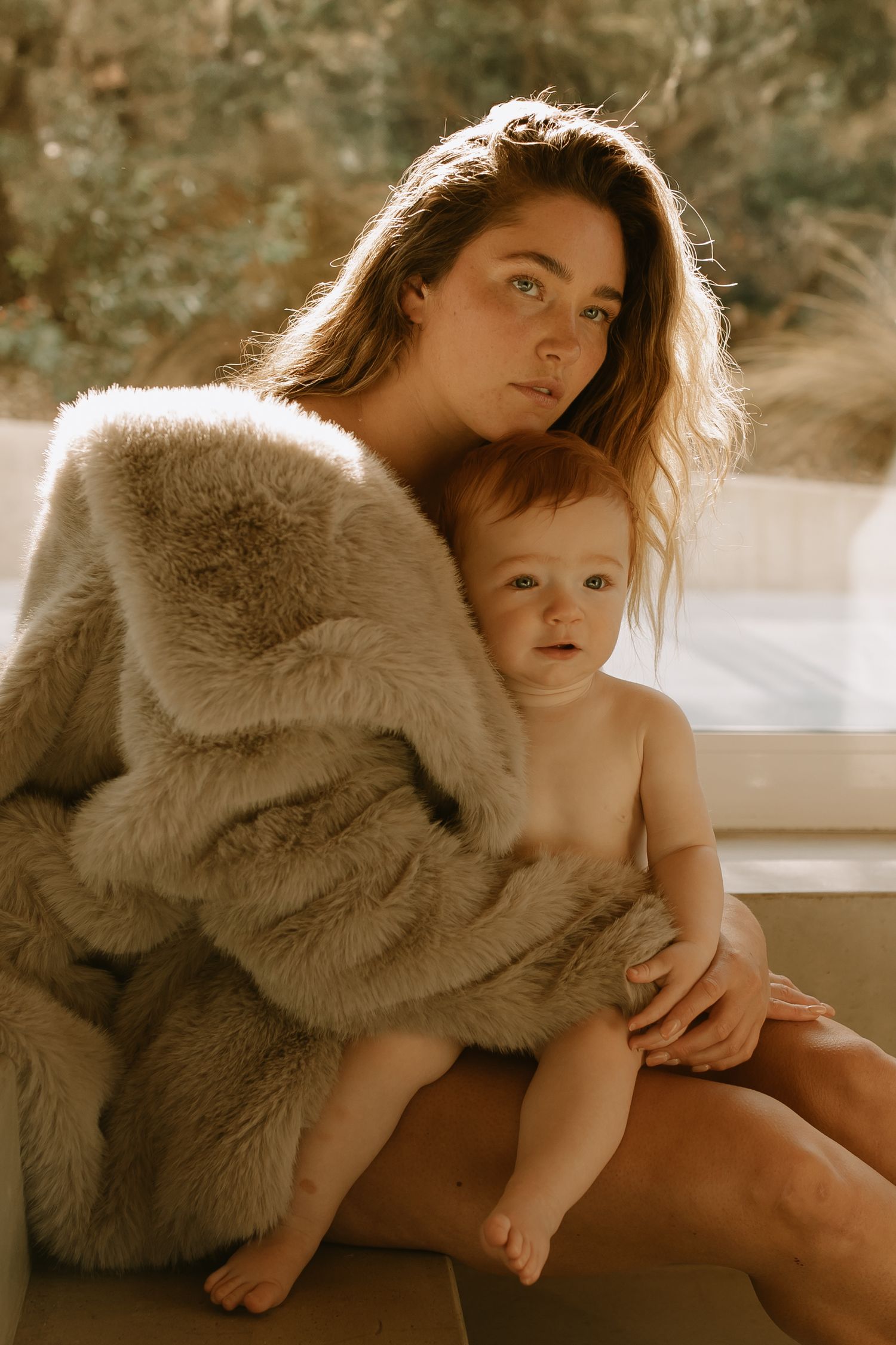 Artistic sepia-toned portrait of parent and child in cozy fur blanket by window with warm backlighting.