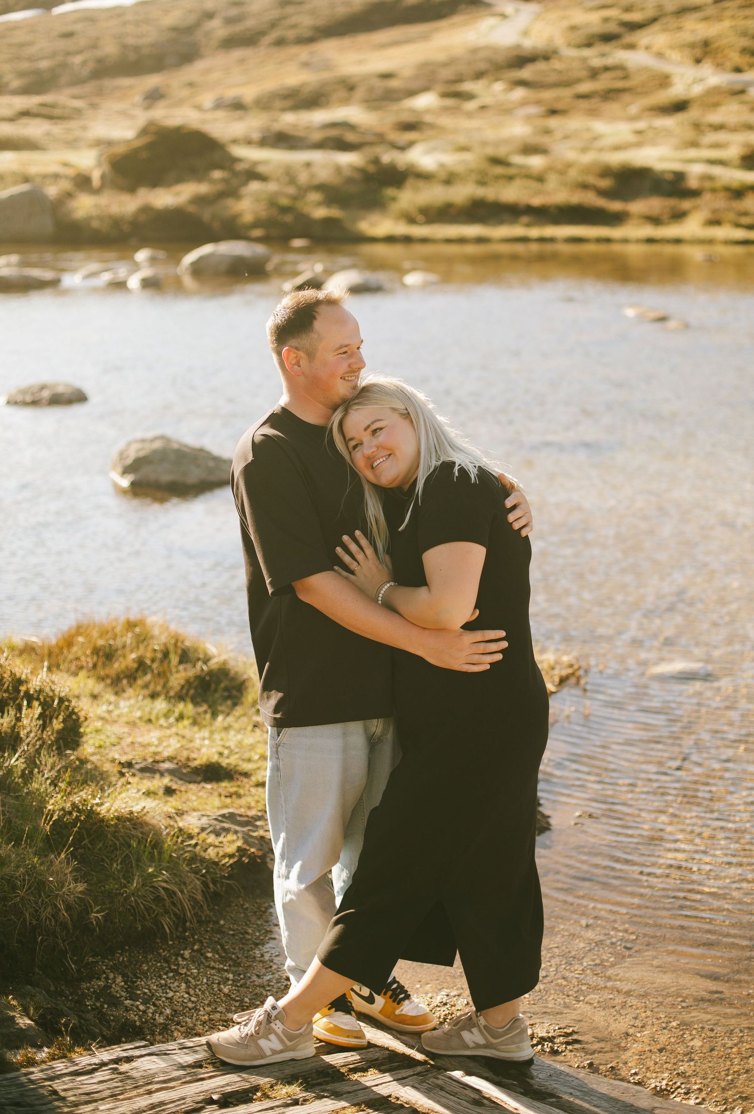 A couple embraces lovingly by a serene lake with rocky shores on a golden afternoon.