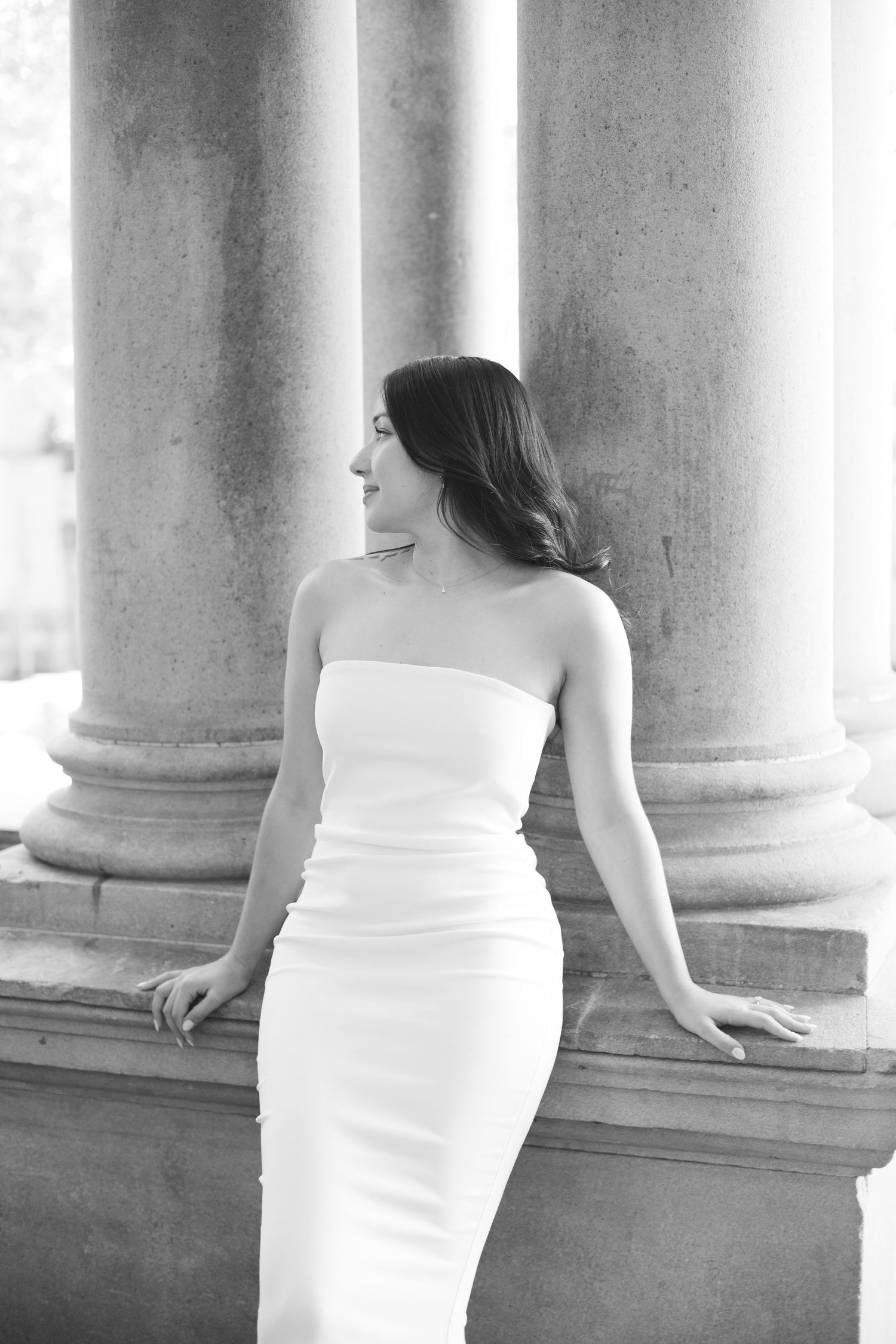 Elegant black and white photo of someone in a strapless white fitted dress leaning against classical stone columns.