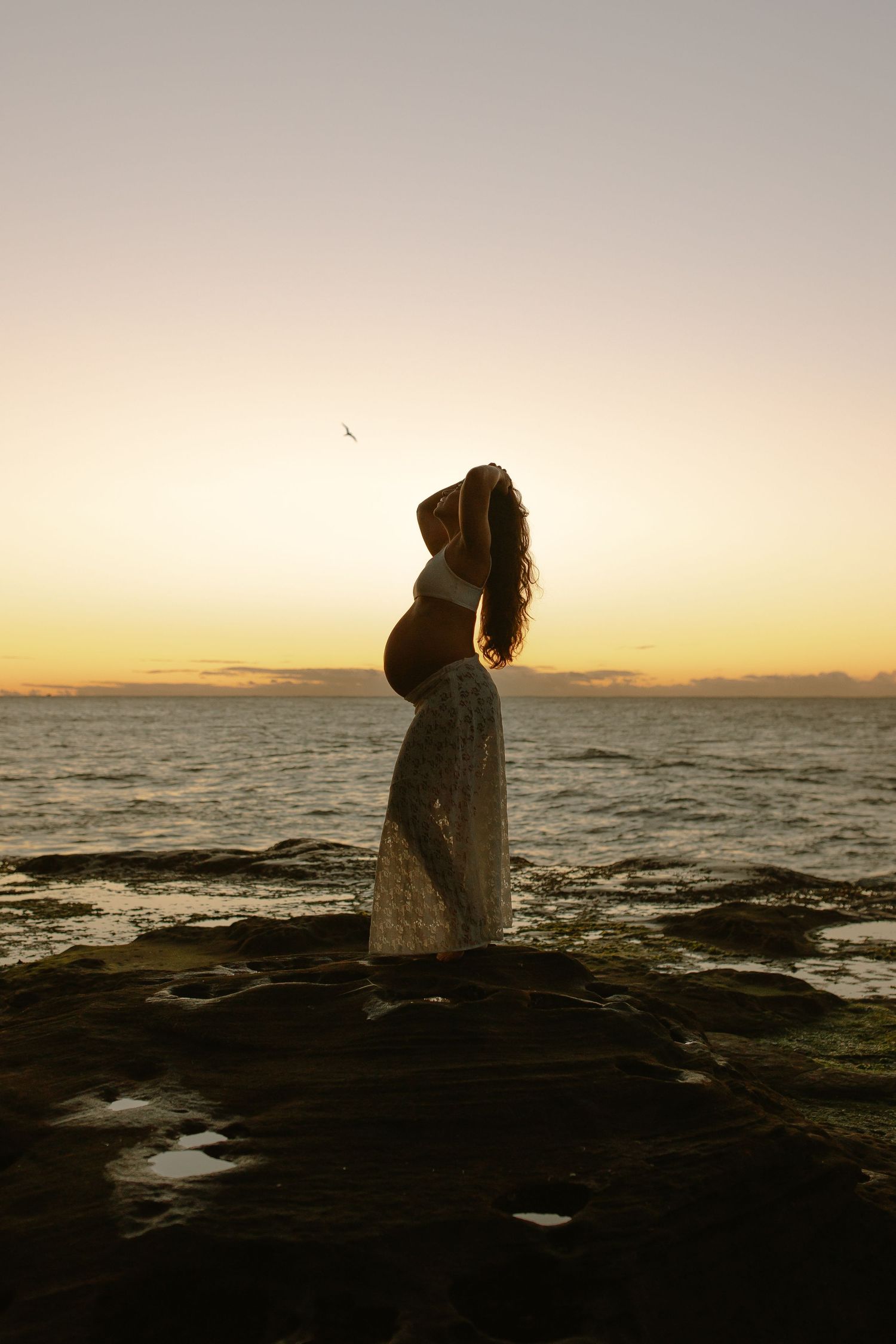 A silhouetted pregnant woman stands on rocky shore against golden sunset sky and ocean backdrop.