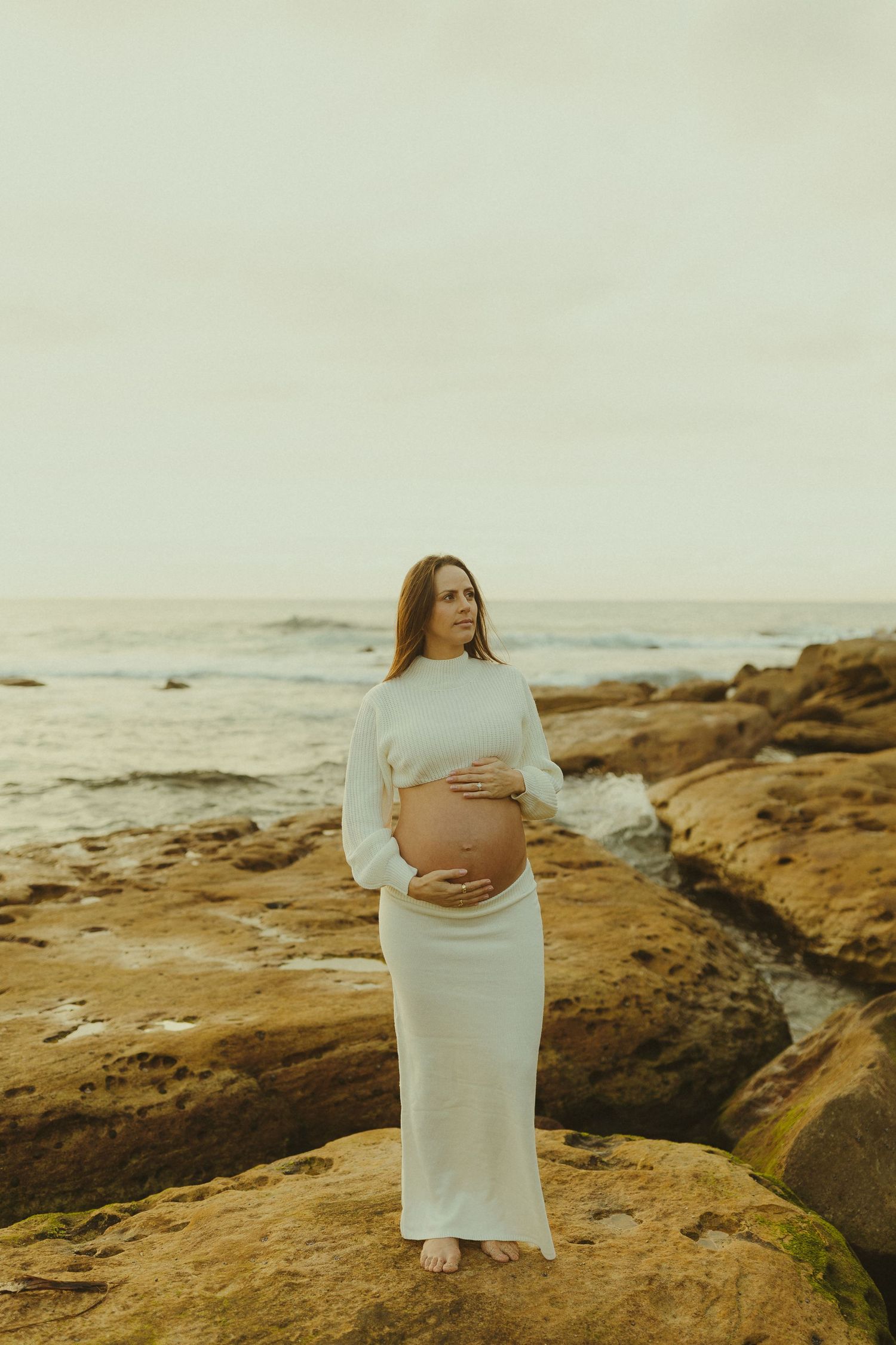 Pregnant woman in white dress poses on rocky coastal outcrop during golden hour maternity shoot.