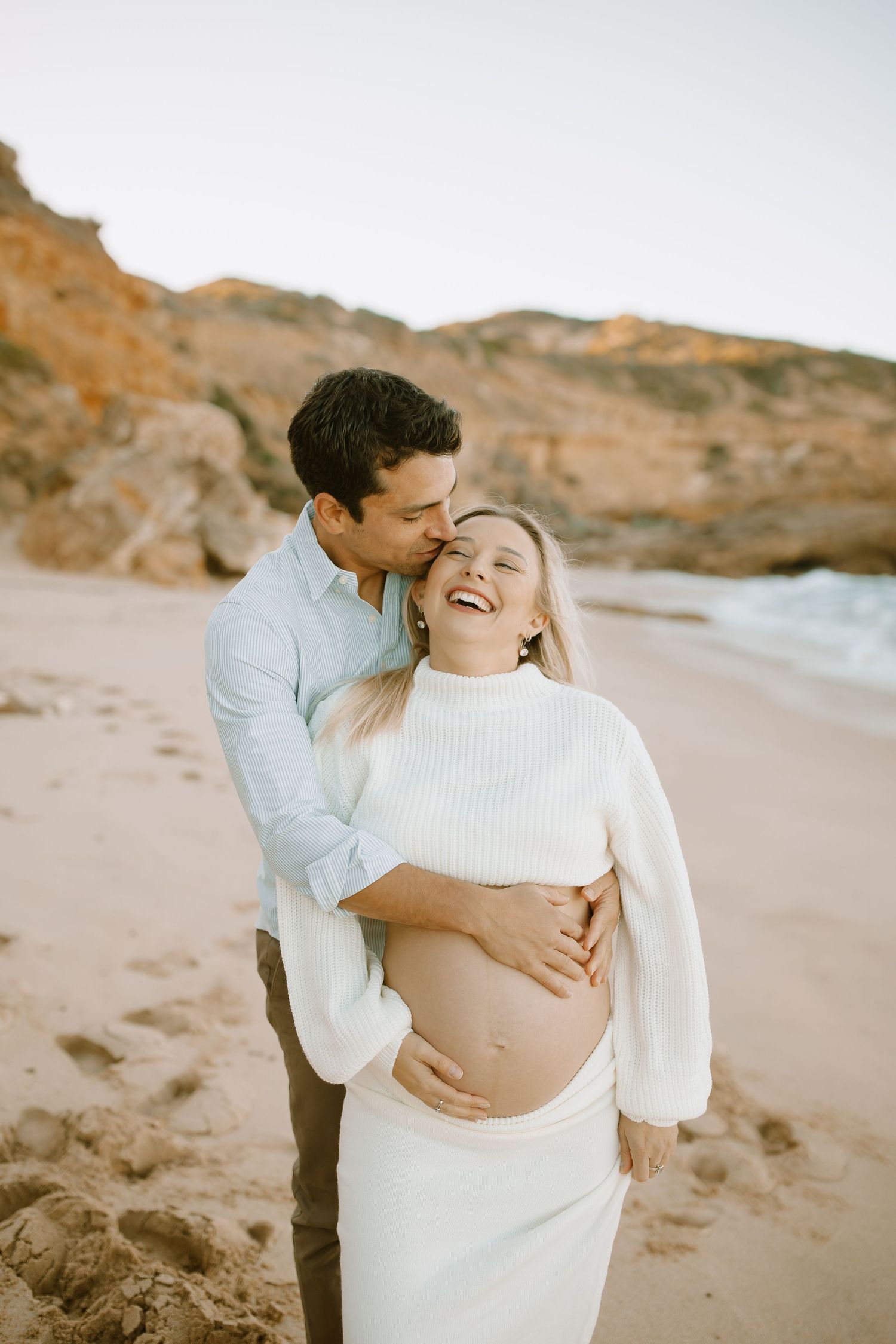 An intimate maternity photo shows a loving couple sharing a tender moment by the ocean at sunset.