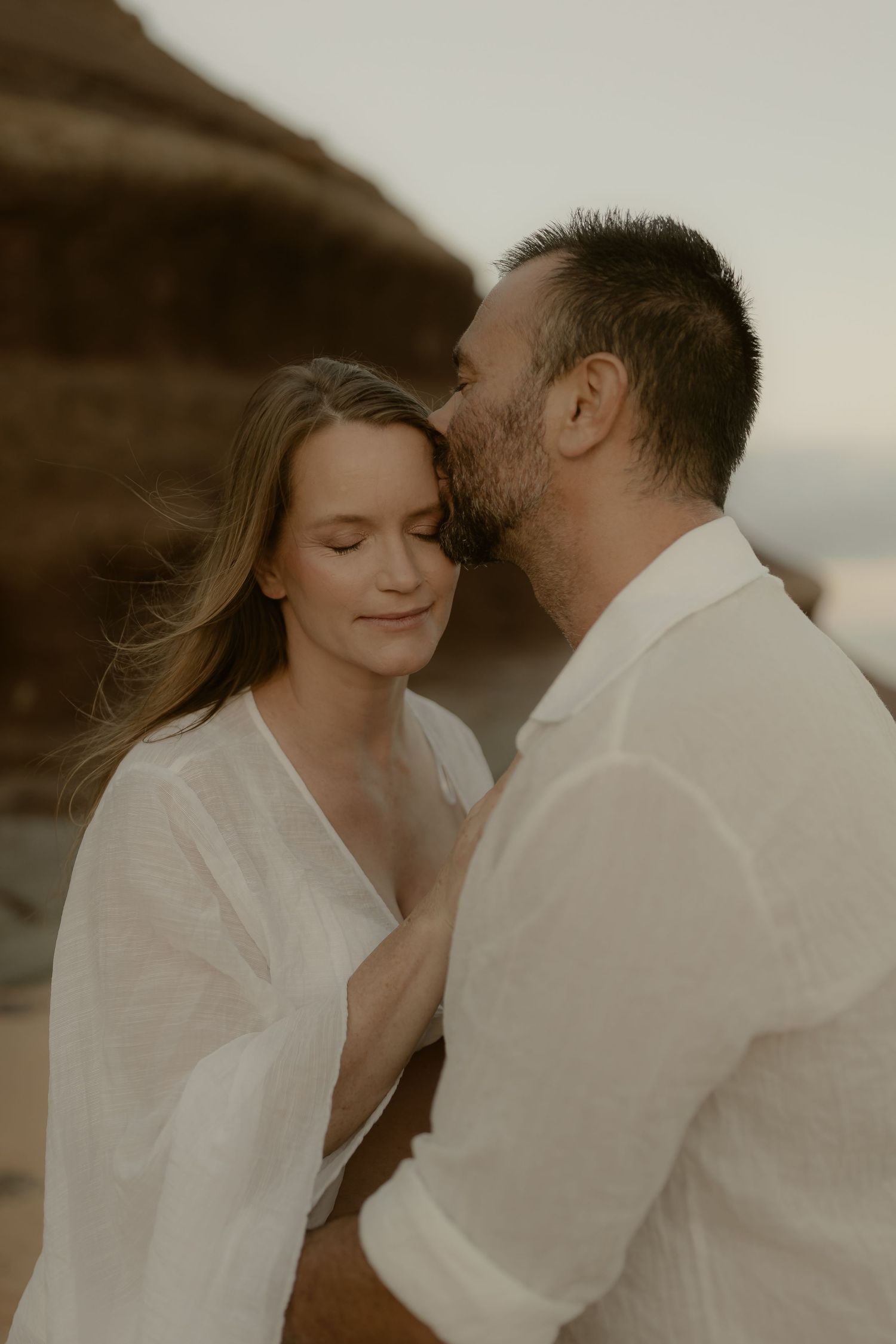 Tender embrace between couple wearing white clothing against natural rocky backdrop.