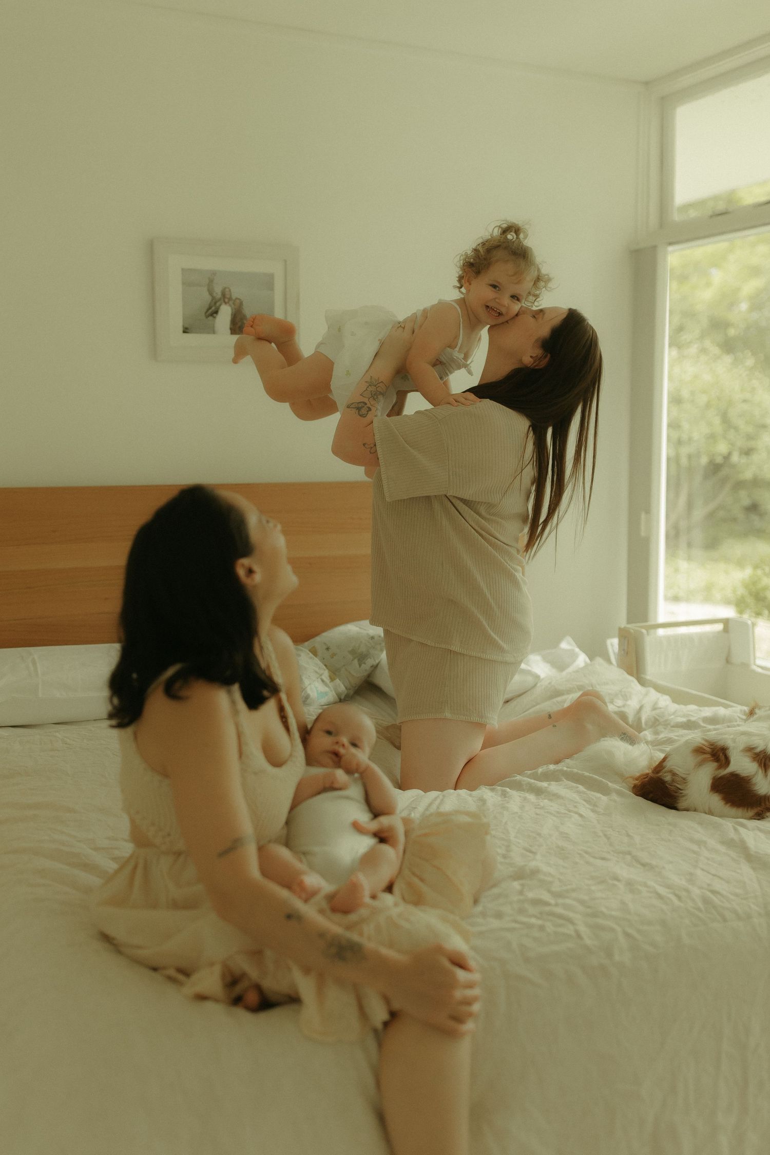A tender family moment captured on a white bed as two adults play with infants in a bright, sunlit room.