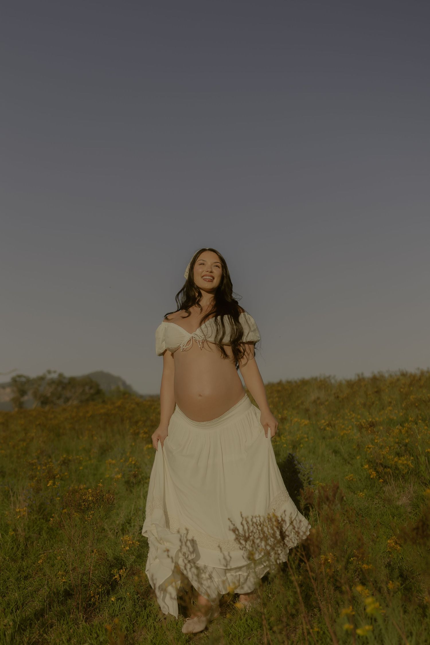 A flowing white dress twirls gracefully in a golden meadow at sunset with mountains in the background.