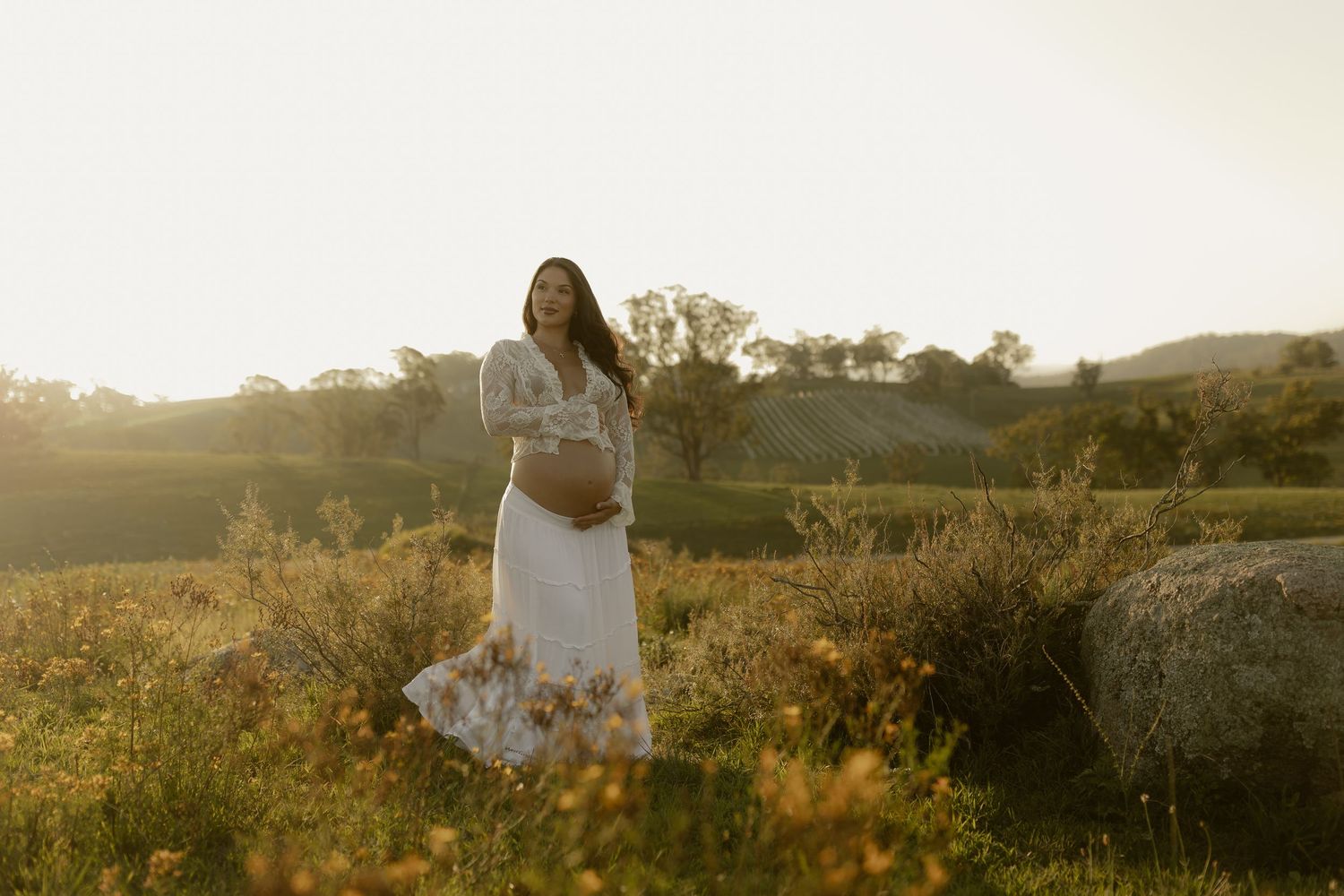 Person in white dress stands peacefully in a golden field at sunset with mountains in background.
