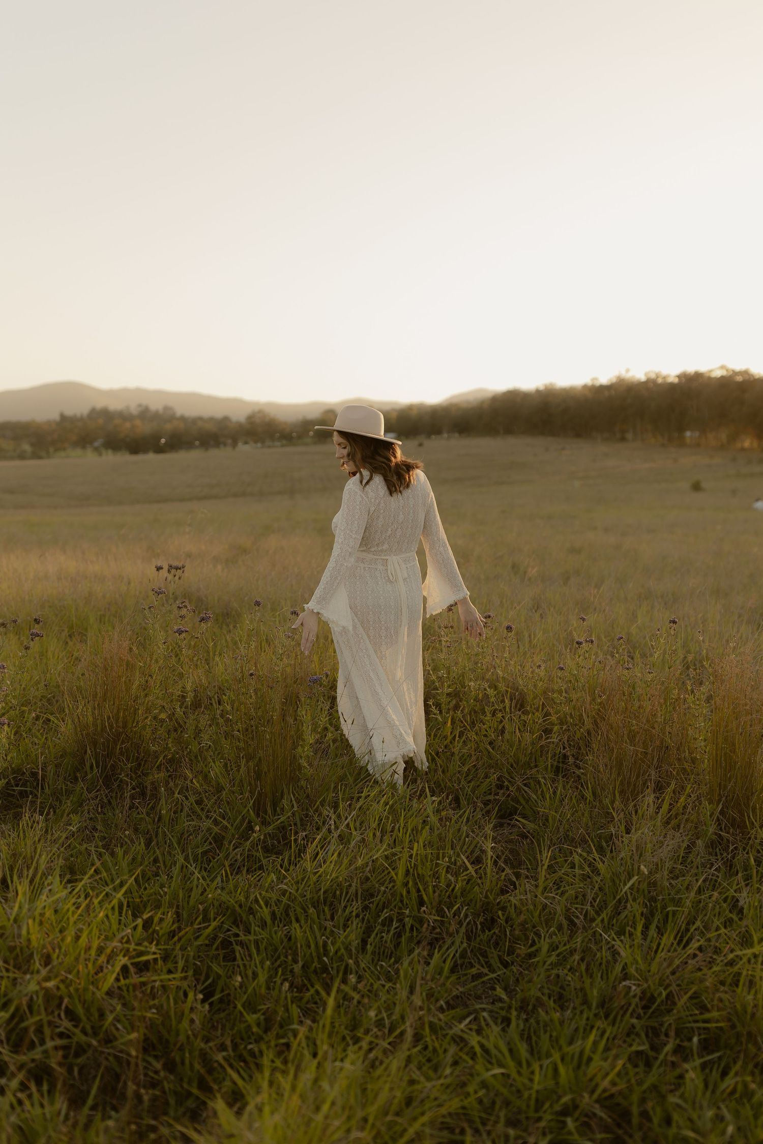 A silhouette stands in a serene meadow at sunset wearing a flowing white dress and wide-brimmed hat.