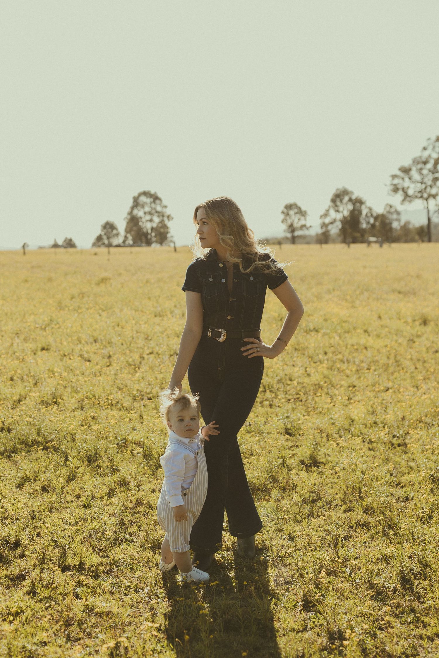Two people stand together in a golden field during sunset with trees in the background.