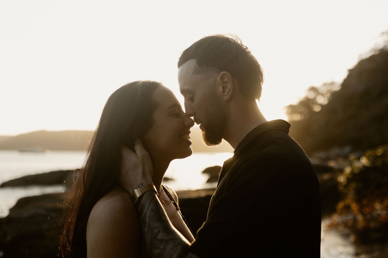 A romantic couple embraces in silhouette against a scenic mountain and lake view during golden hour sunset.