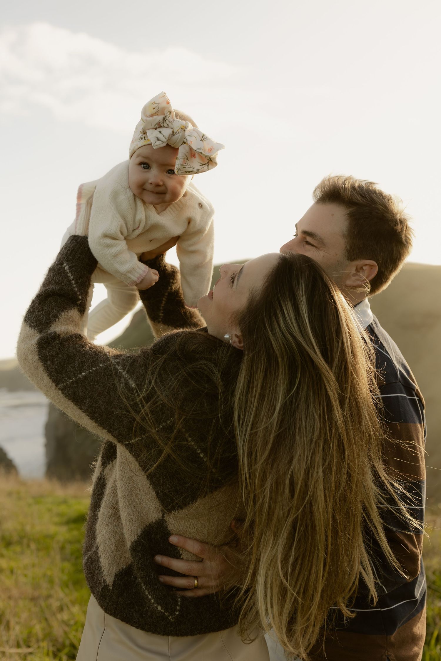Young couple holding smiling baby outdoors near coastal cliffs, mother lifting infant in floral headband.