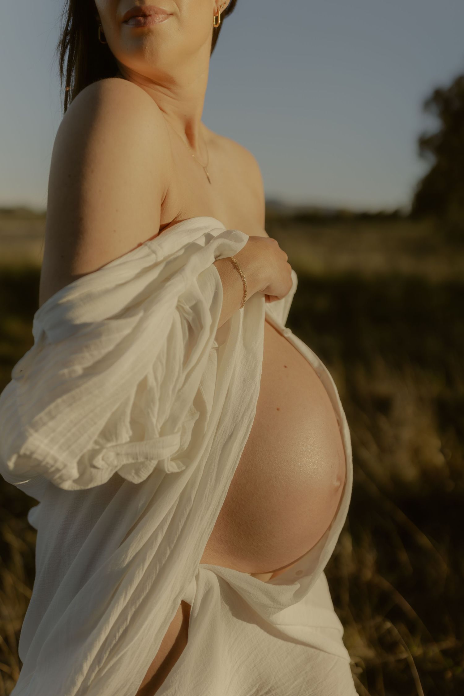 Pregnant woman in white muslin wrap exposing baby bump in golden field at sunset.