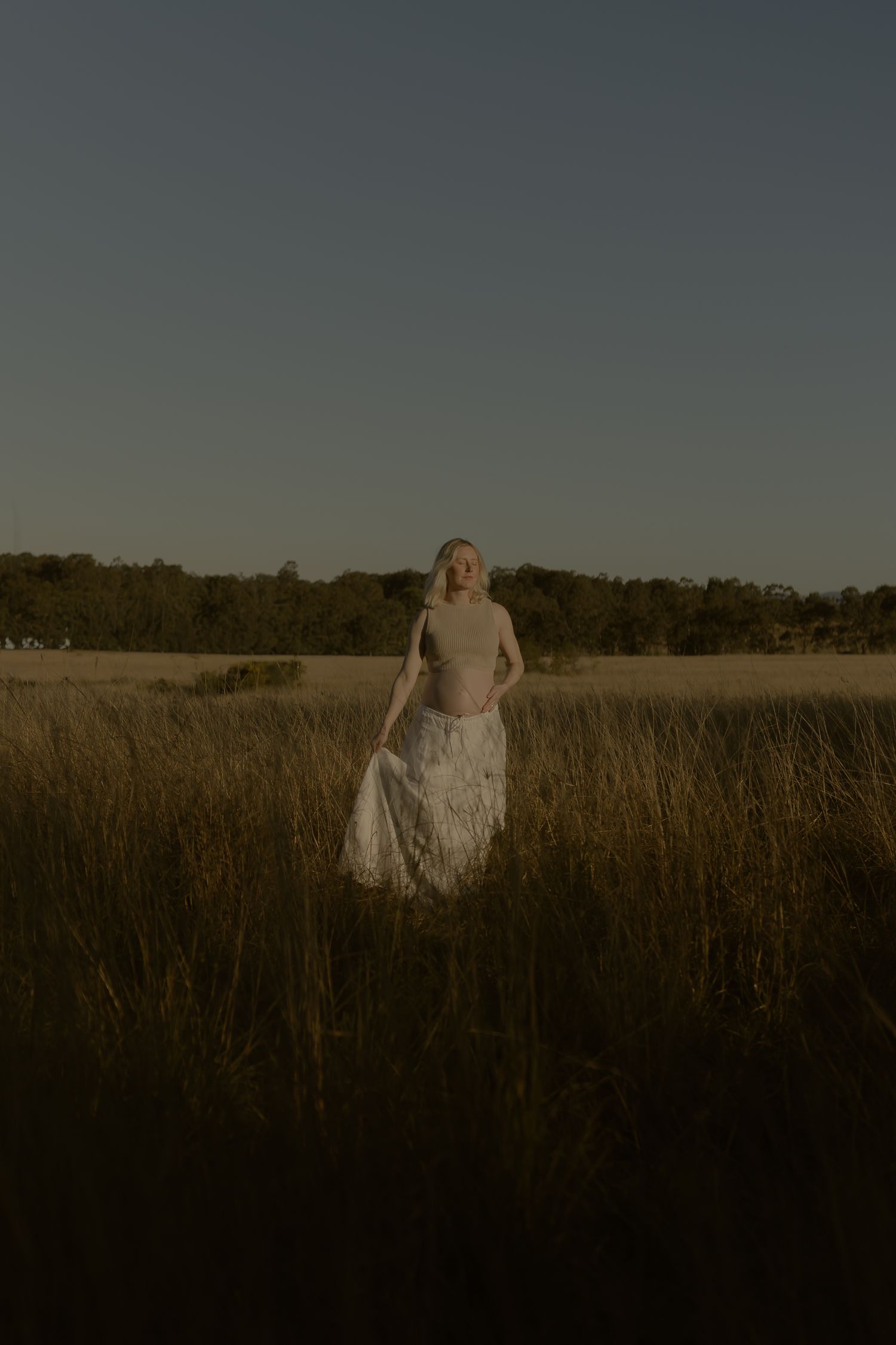 Woman in white skirt standing in golden grass field at dusk with trees in background.