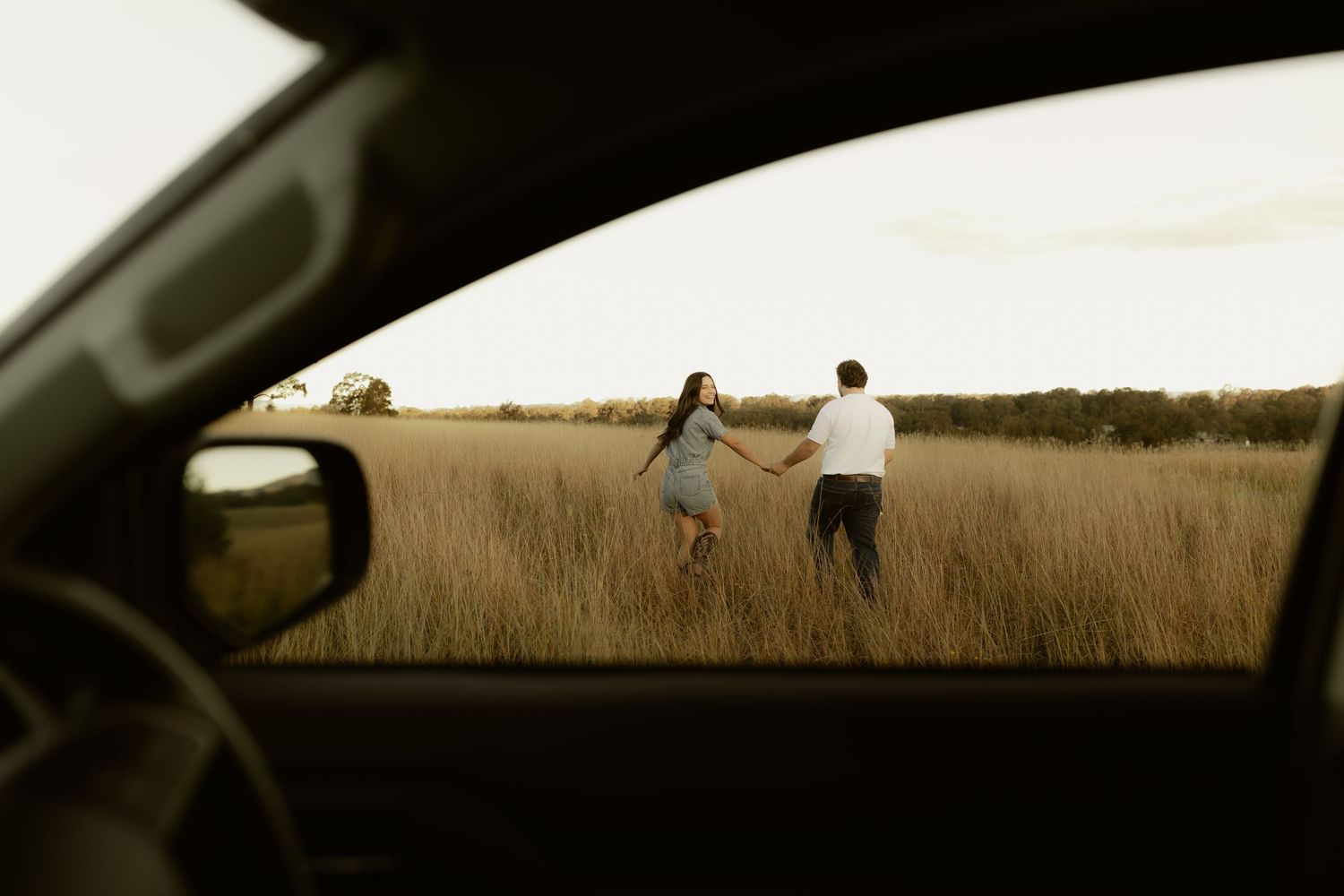 Couple holding hands running through golden field, photographed through car window at sunset.