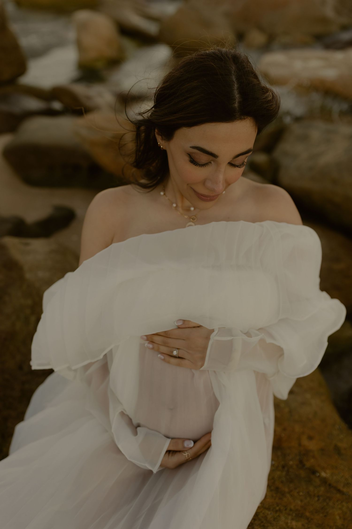 Pregnant woman in white off-shoulder gown cradling baby bump, sitting among rocks in elegant maternity photo.