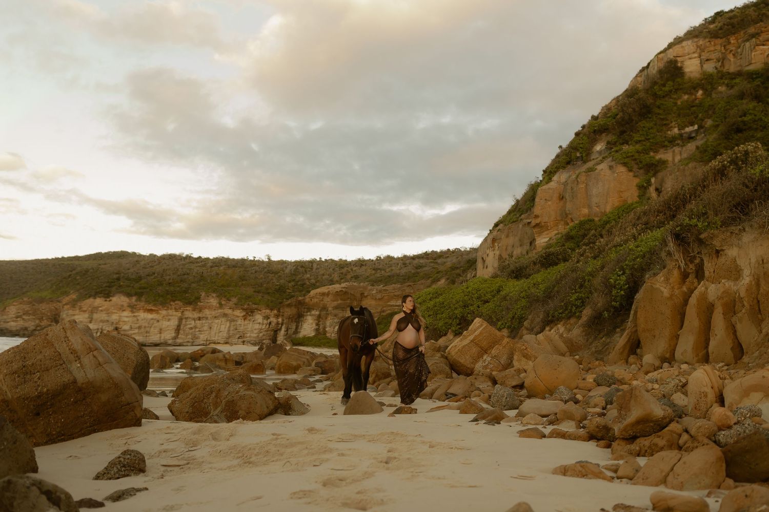 Two people walking on a rocky beach with sandstone cliffs and lush greenery under a cloudy sky.