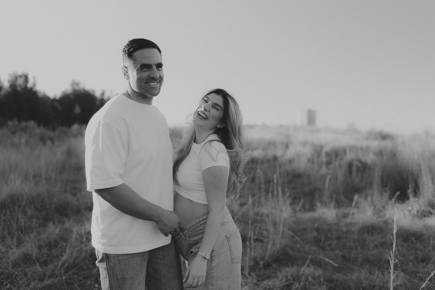 Black and white photo of a smiling pregnant couple holding hands in a field at sunset.