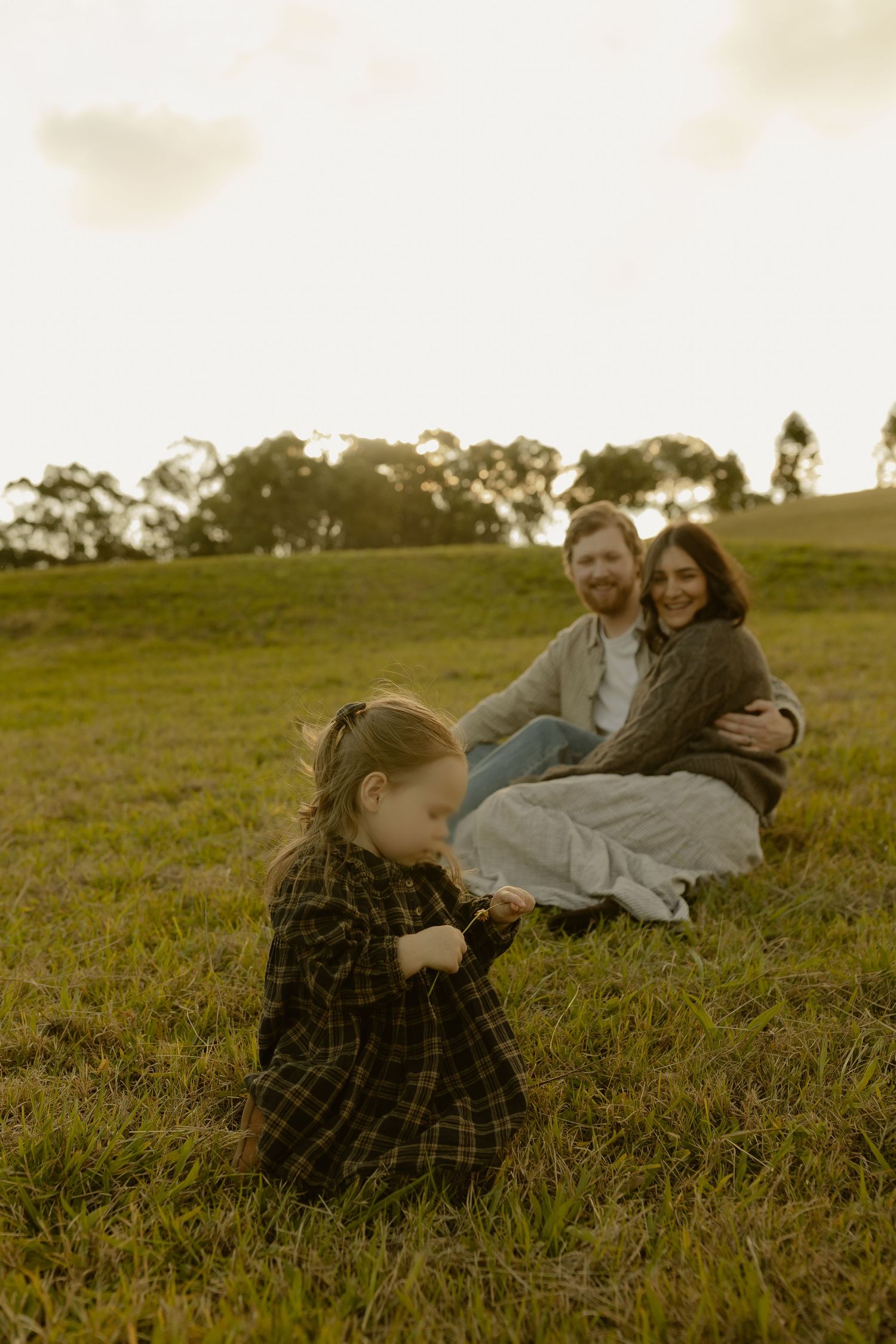 A young girl in a plaid dress kneels in the grass while her parents sit together smiling on a hill.