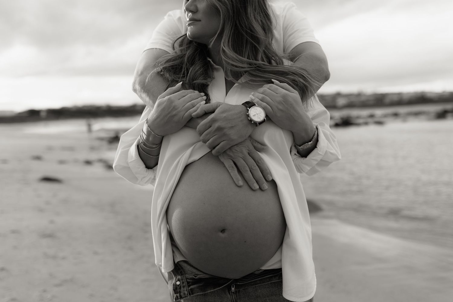 Black and white photo of pregnant woman outdoors, partner embracing her from behind, both hands cradling bare baby bump.