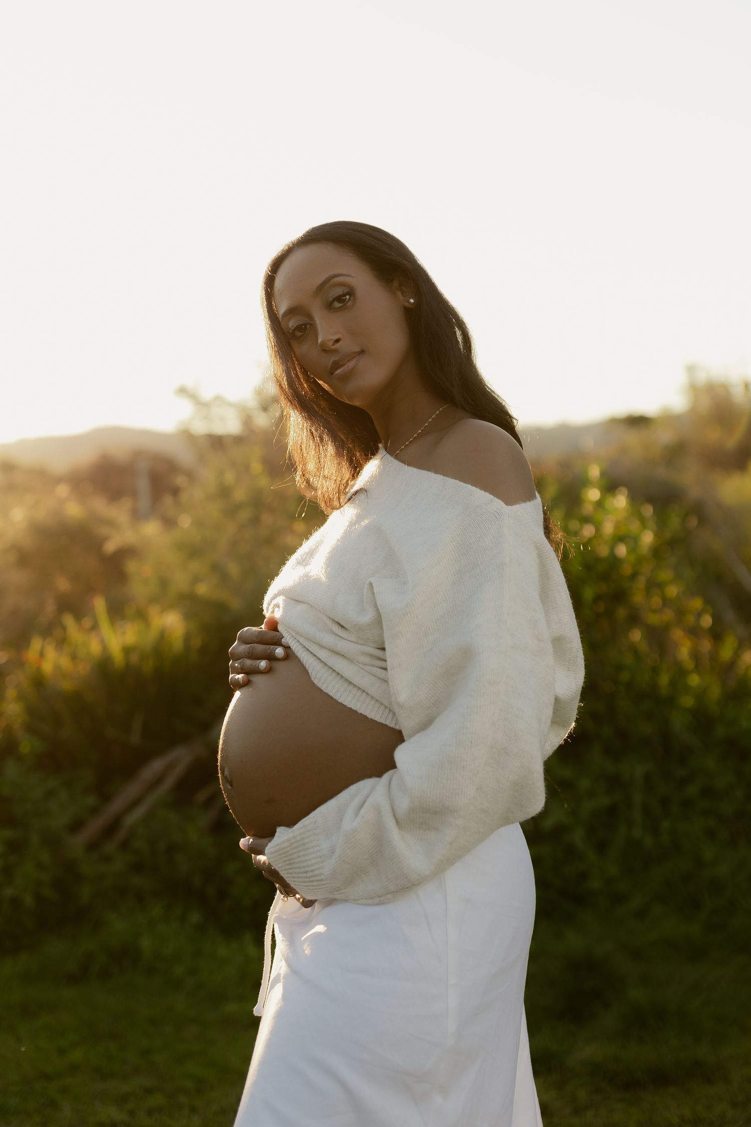 Pregnant woman in white off-shoulder sweater cradling baby bump outdoors at golden hour sunset.